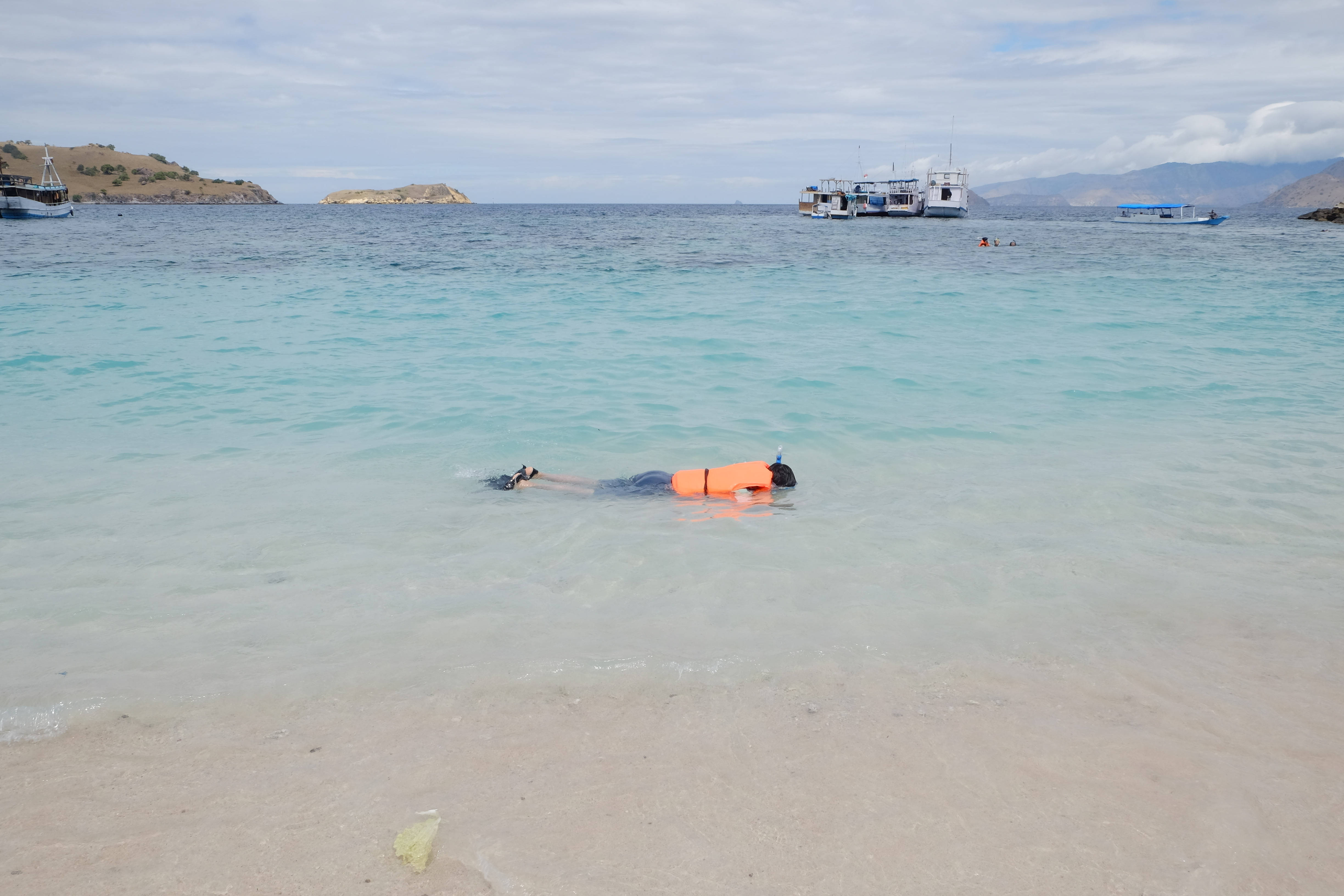 Snorkeling 桌布 壁紙 — 一個人在清澈的藍綠色水中仰面漂浮，靠近沙灘，有船隻和遠處的島嶼，天空多雲。
