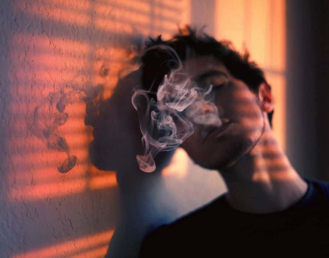bad 桌布 壁紙 — 一個人在溫暖的金色光線中呼出煙霧，光線穿過百葉窗，在紋理牆壁上投下戲劇性的陰影。