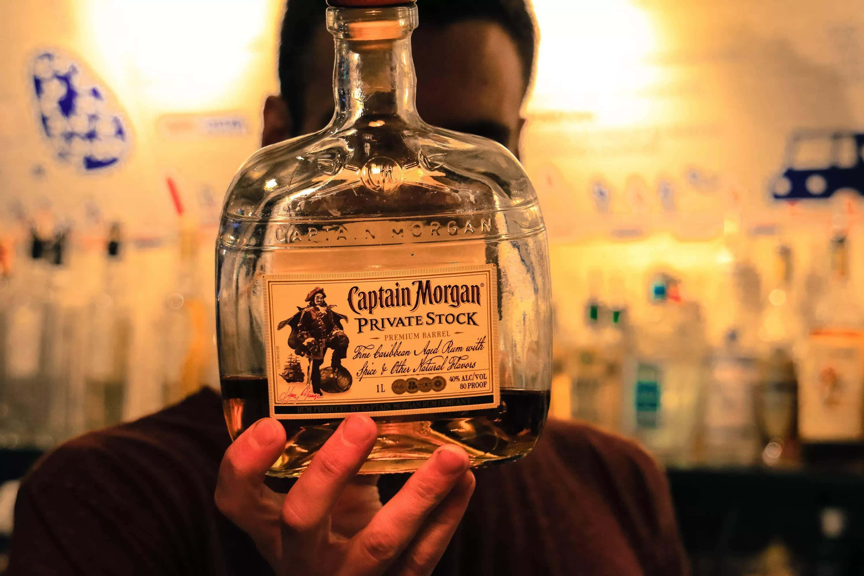 alcohol 桌布 壁紙 — 一個人在燈光溫暖的酒吧裡，手持一瓶Captain Morgan Private Stock蘭姆酒，身後是模糊的酒架。