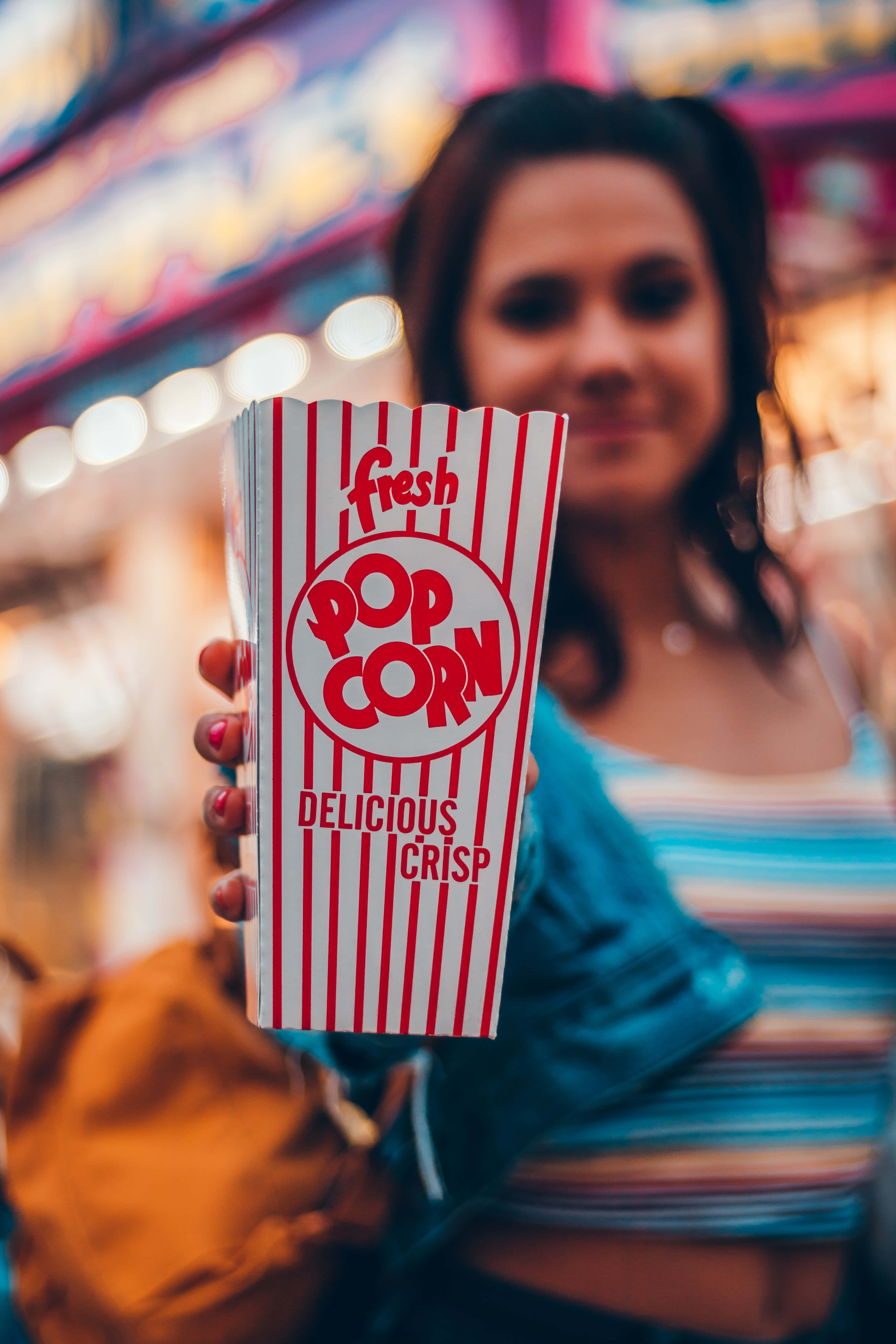 Popcorn 桌布 壁紙 — 一個人在燈火通明的嘉年華會上，手持紅白條紋的爆米花盒，背景是模糊的彩色燈光。