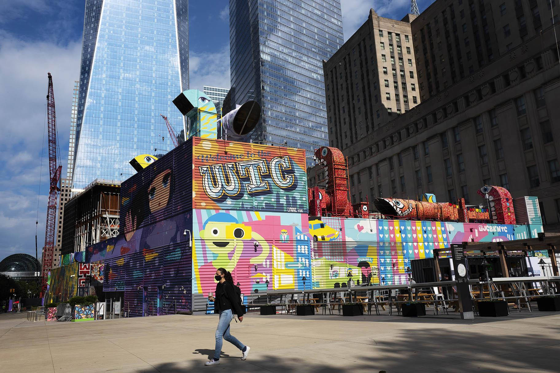 New York Aesthetic 桌布 壁紙 — 一個人在現代城市廣場走過色彩繽紛的壁畫裝置，廣場上有玻璃摩天大樓和建築起重機，天空部分多雲。
