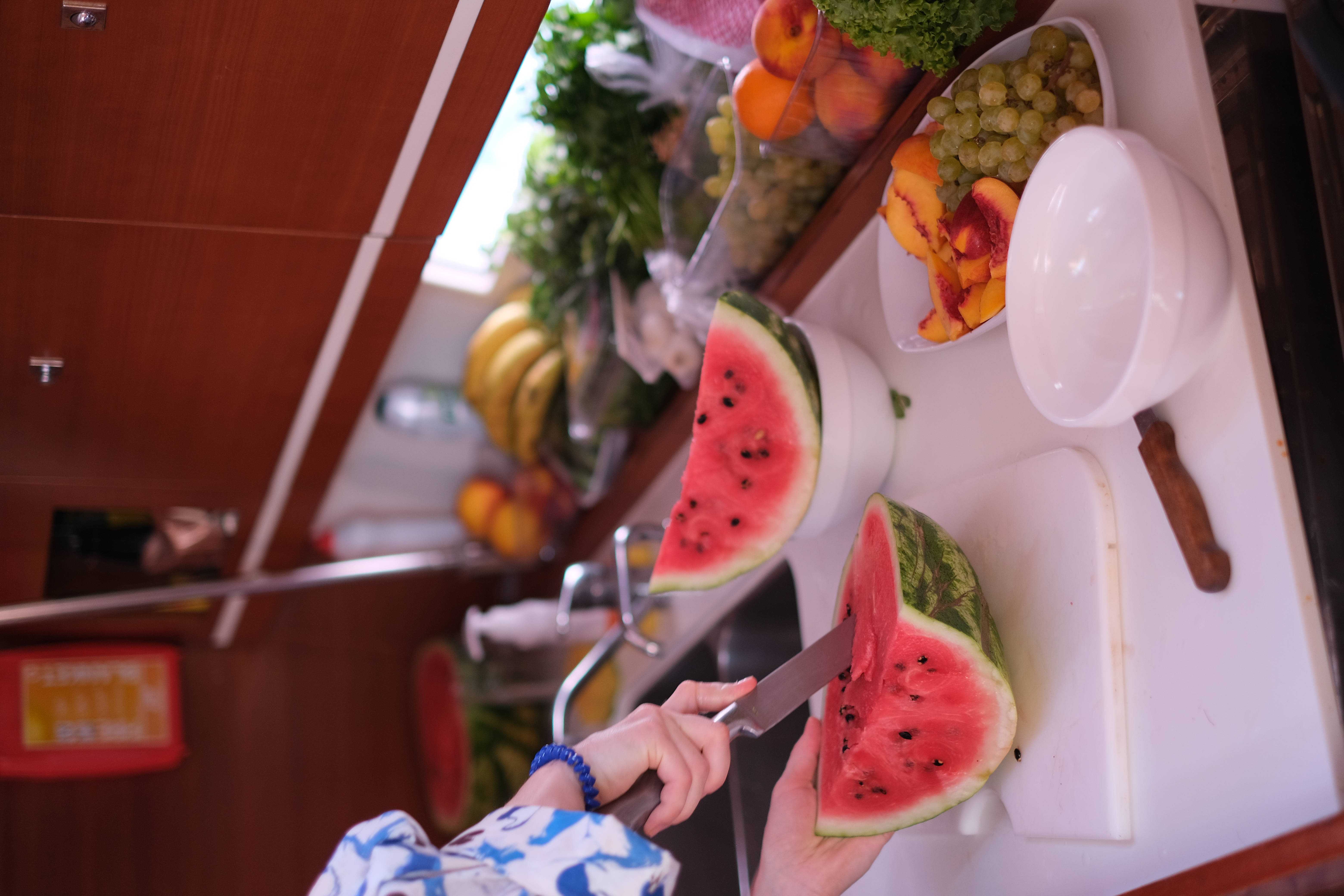 Watermelon 桌布 壁紙 — 一個人在白色砧板上切西瓜，在一個緊湊的木製廚房裡，周圍擺滿了新鮮水果。