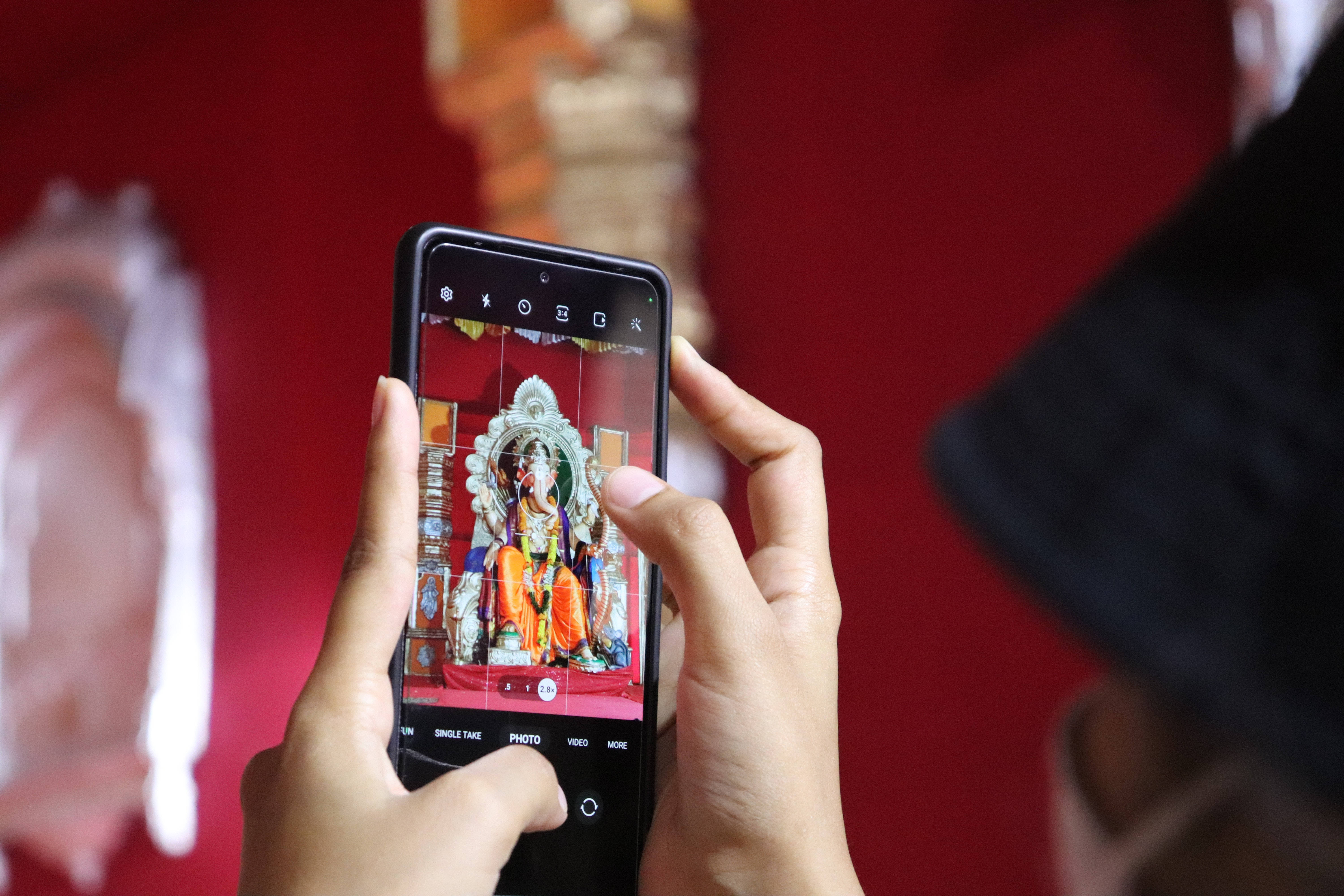 ganesh 4K 桌布 壁紙 — 一個人在紅色牆壁的寺廟裡，用智慧型手機拍攝一個色彩繽紛、裝飾華麗的神像。