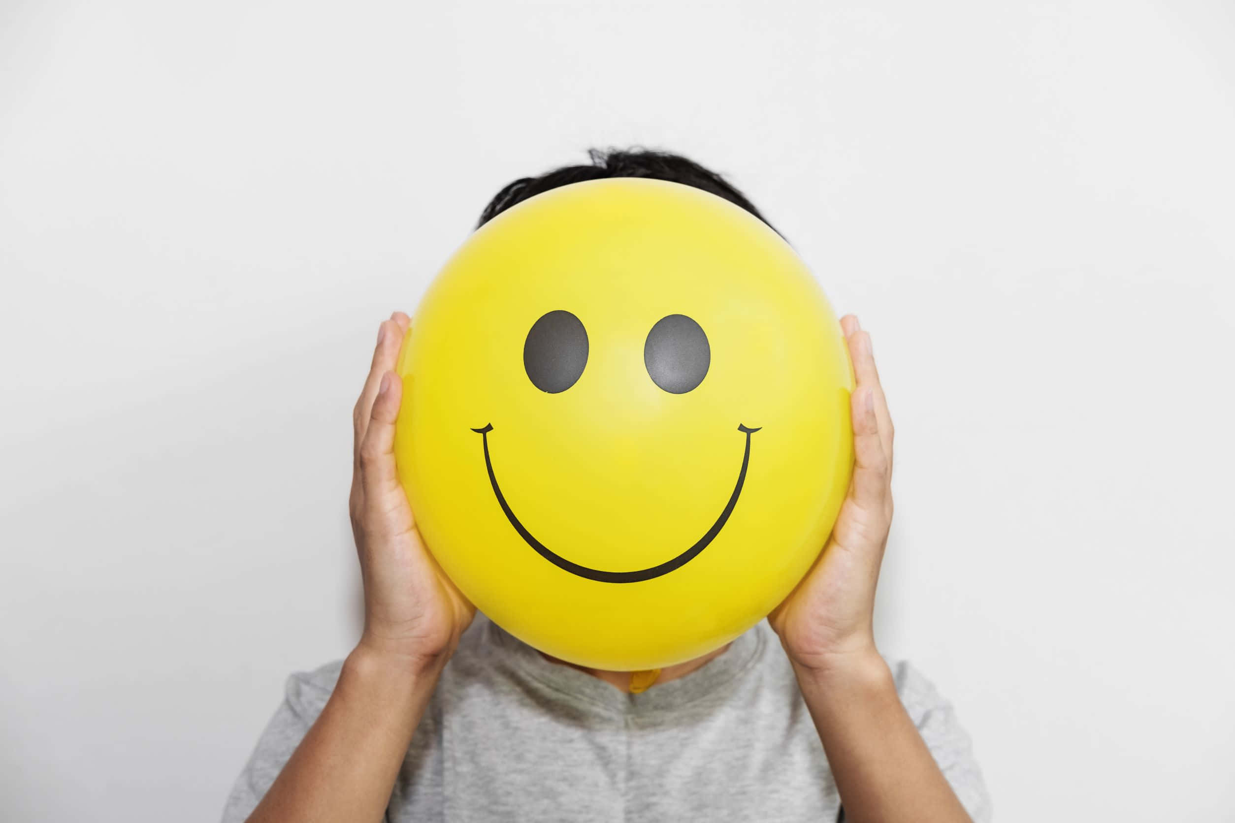 positivity 桌布 壁紙 — 一個人在純白色背景前，將一個亮黃色笑臉氣球舉在臉上。