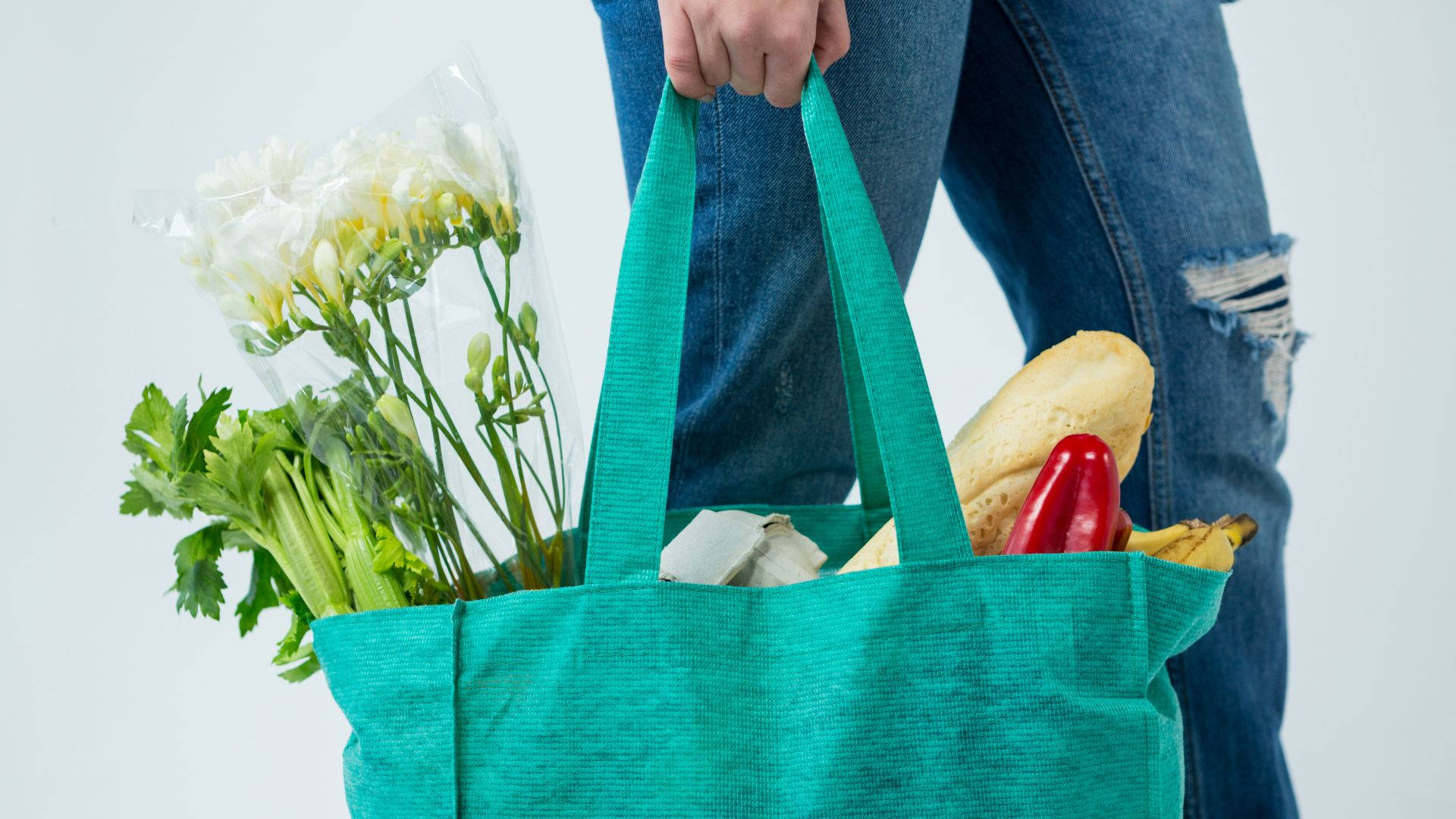 Fruits And Vegetables 桌布 壁紙 — 一個人在純白色背景前，拿著一個裝滿新鮮雜貨和白色花朵的藍綠色可重複使用購物袋。