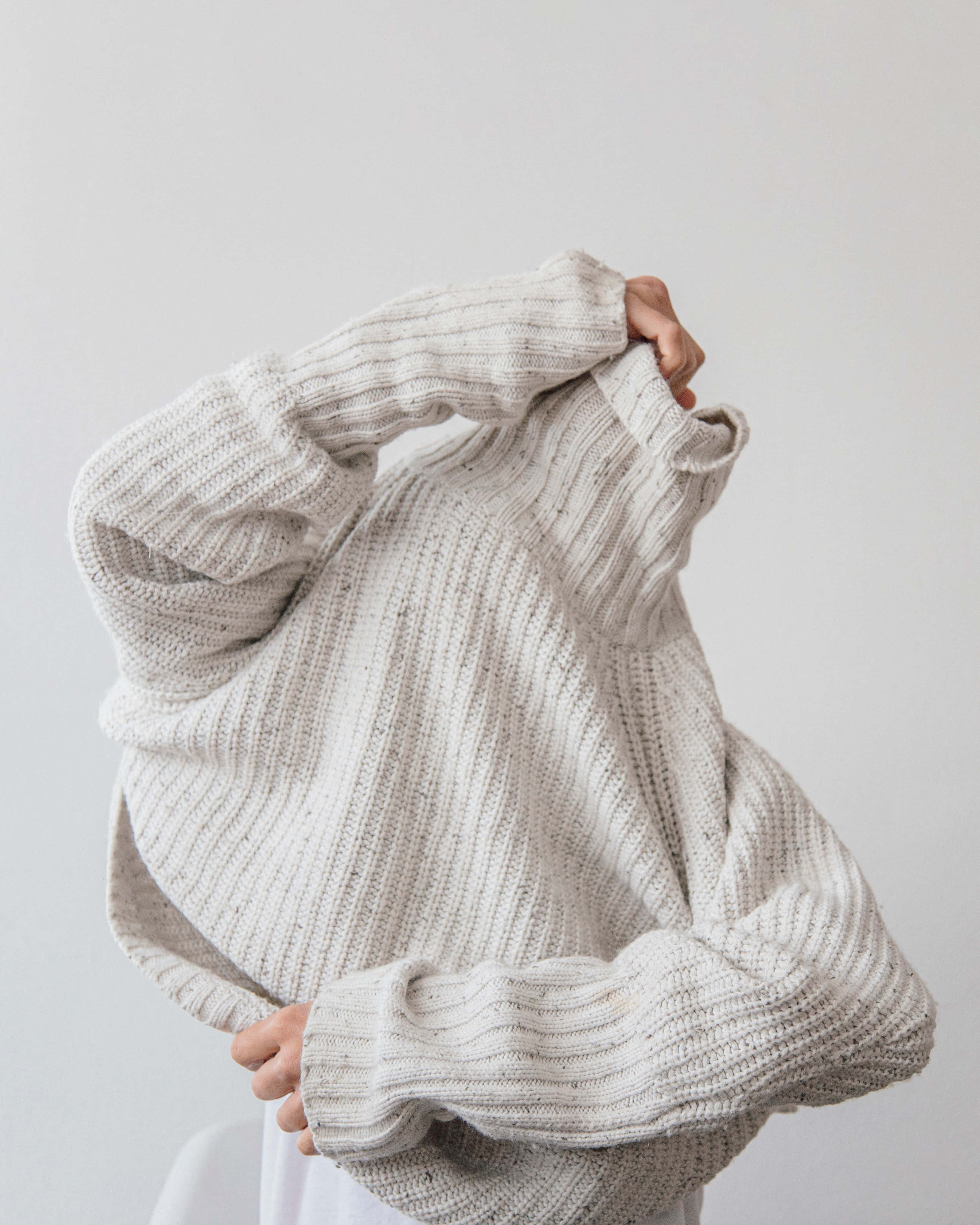 clothing 桌布 壁紙 — 一個人在純白色背景前舉起一件舒適的米色羅紋針織毛衣。