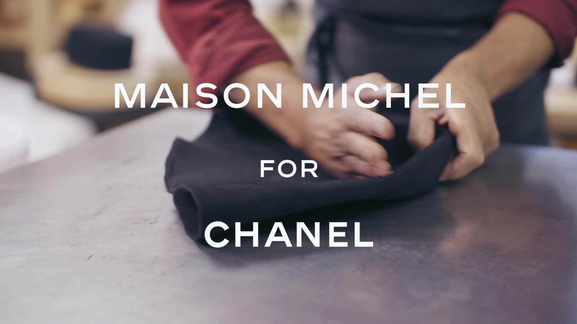 標誌 桌布 壁紙 — 一個人在金屬桌上摺疊黑色布料，帶有 'MAISON MICHEL FOR CHANEL' 文字疊加。