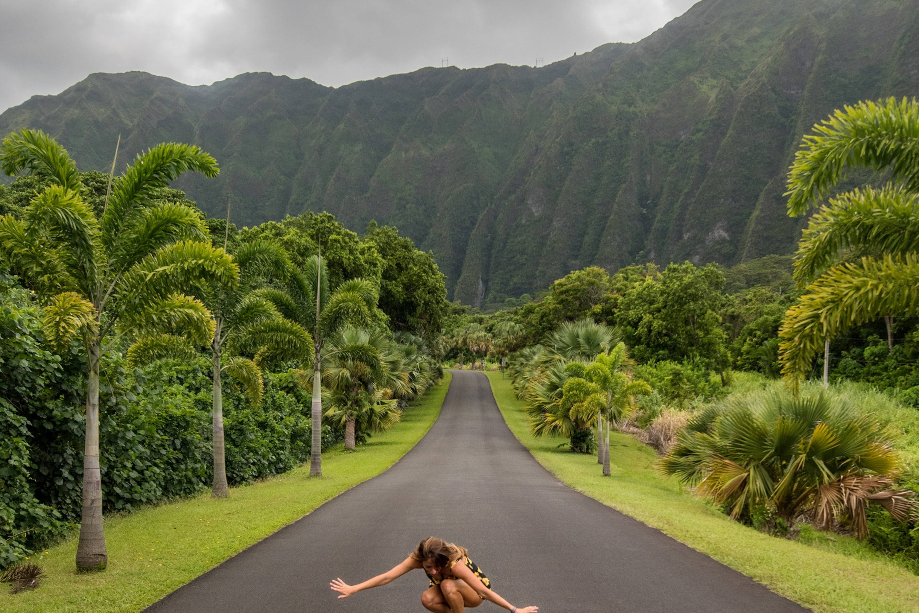 Road 桌布 壁紙 — 一個人在鋪砌的道路上蹲在半空中，兩旁是棕櫚樹和鬱鬱蔥蔥的綠色山脈，天空多雲。