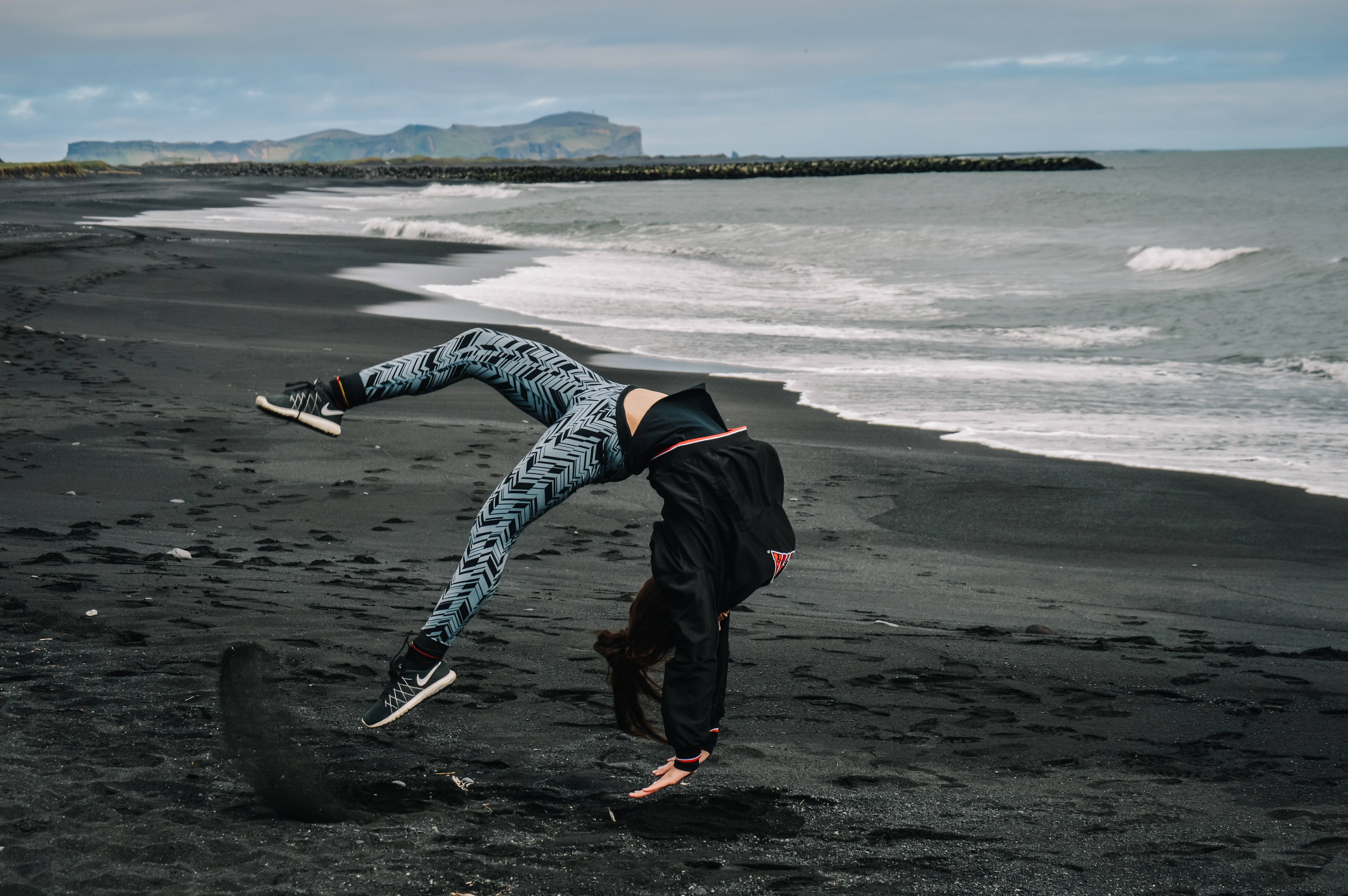 體操 桌布 壁紙 — 一個人在陰天的黑色火山沙灘上，在拍岸的海浪和遠處的懸崖背景下，做一個動態的倒立動作。