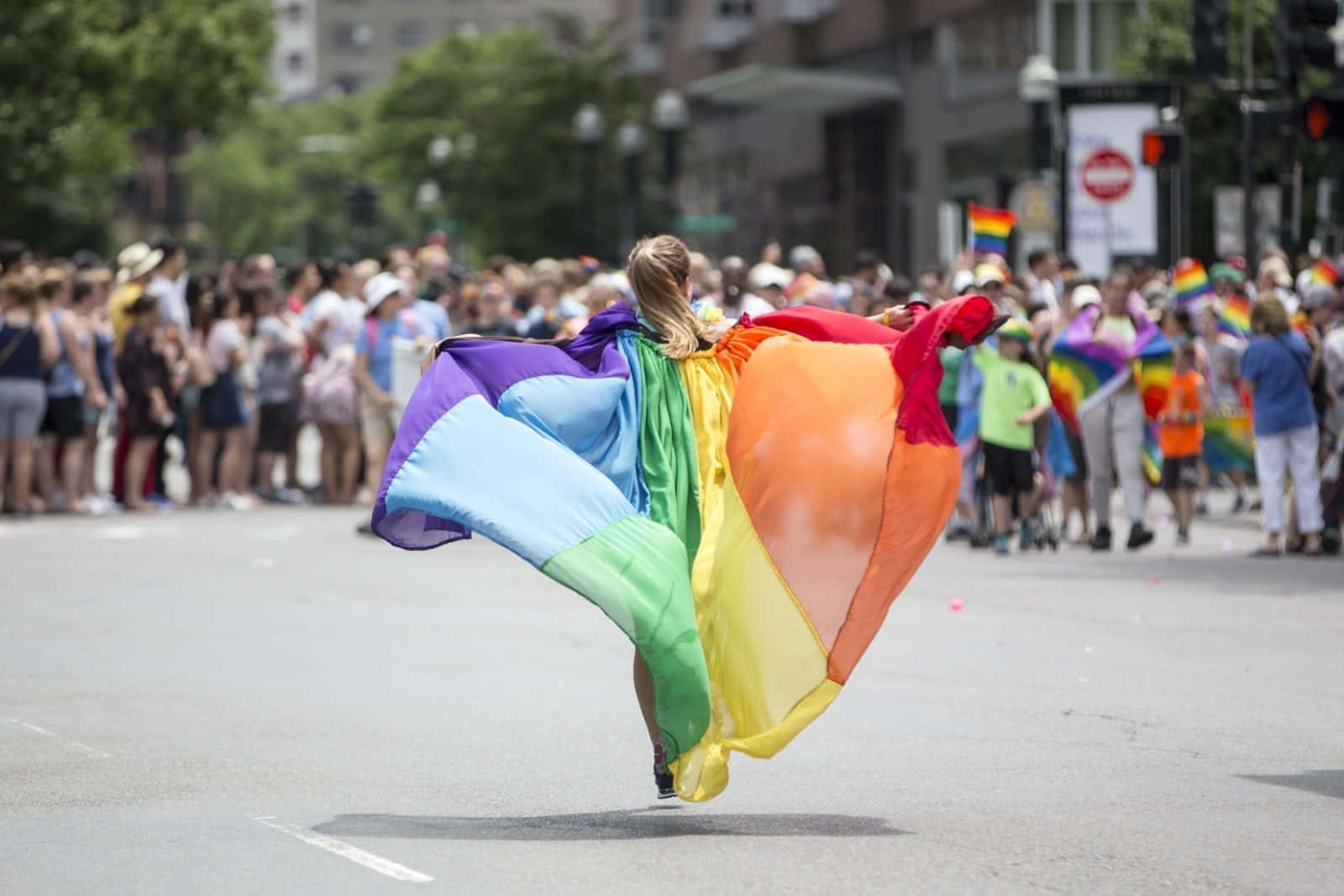 Pride Flag 桌布 壁紙 — 一個人在陽光明媚的城市街道上奔跑，在驕傲遊行中揮舞著一面巨大的彩虹旗，人行道上的群眾歡呼雀躍。