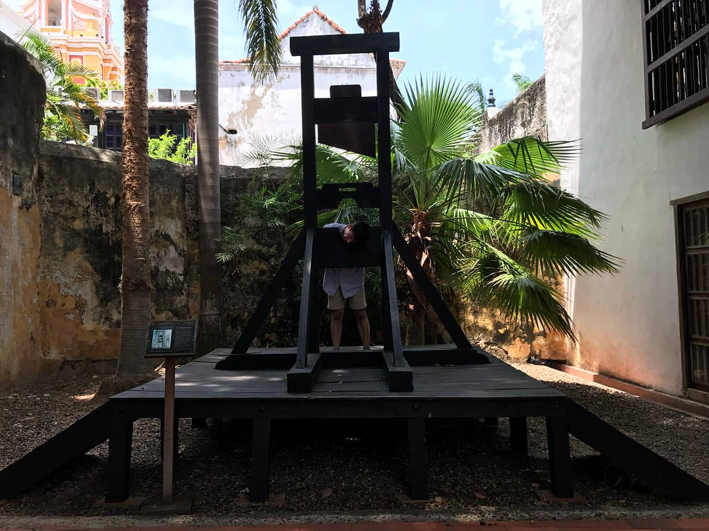 Museum 桌布 壁紙 — 一個人在陽光明媚的庭院中，在棕櫚樹和舊牆環繞下，於一個大型黑色斷頭台複製品內頑皮地擺姿勢。
