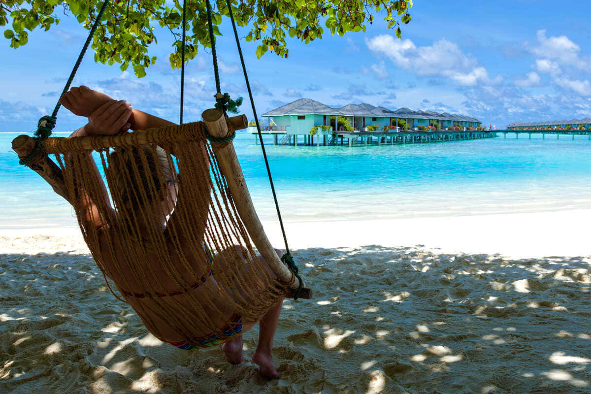 Jamaican Beach 桌布 壁紙 — 一個人在陽光明媚的海灘上，在編織吊床上放鬆，遠處是碧綠的海水和水上平房。