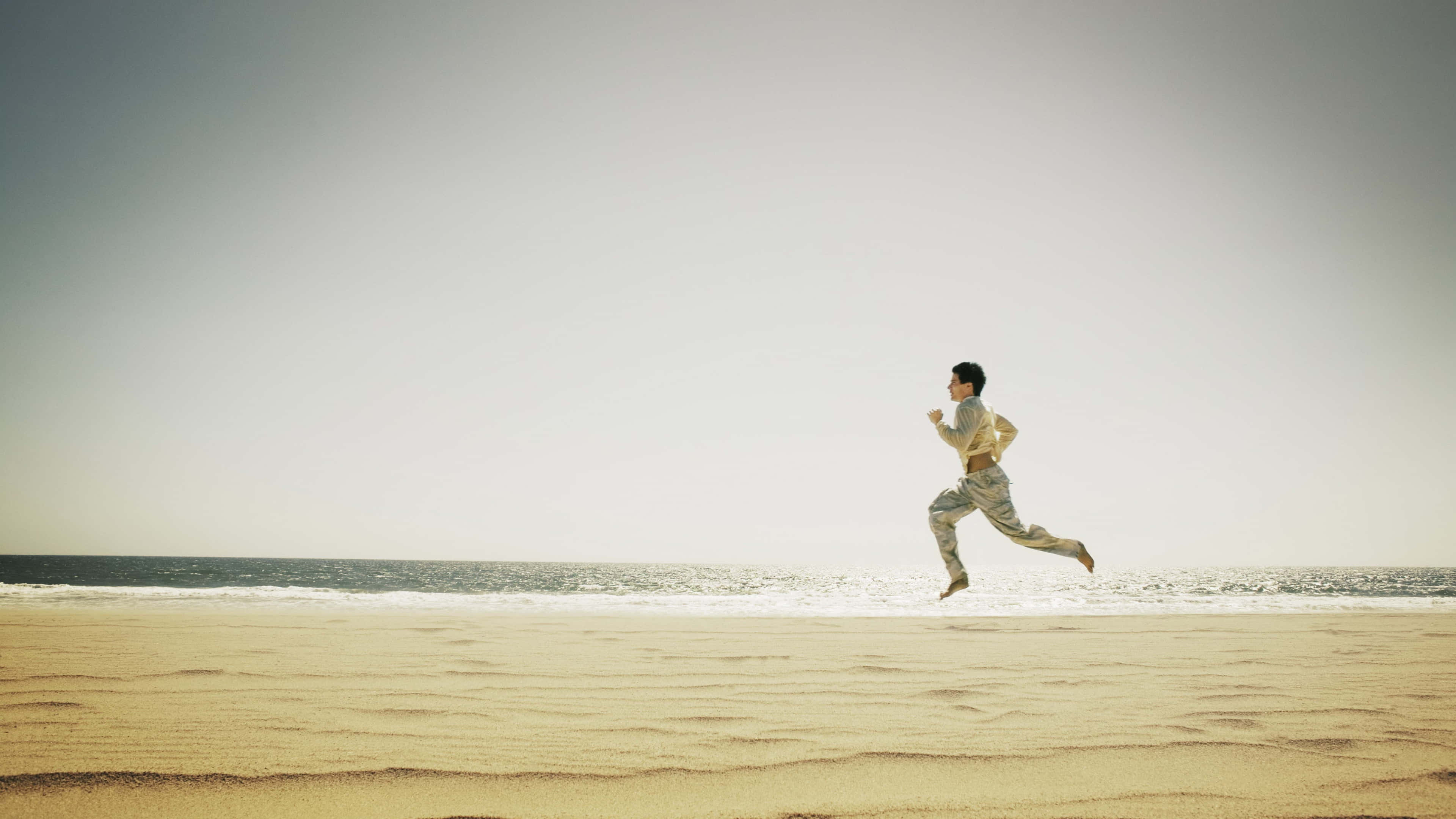 Running 桌布 壁紙 — 一個人在陽光普照的沙灘上快樂地奔跑，海浪在蒼白的天空下翻滾。