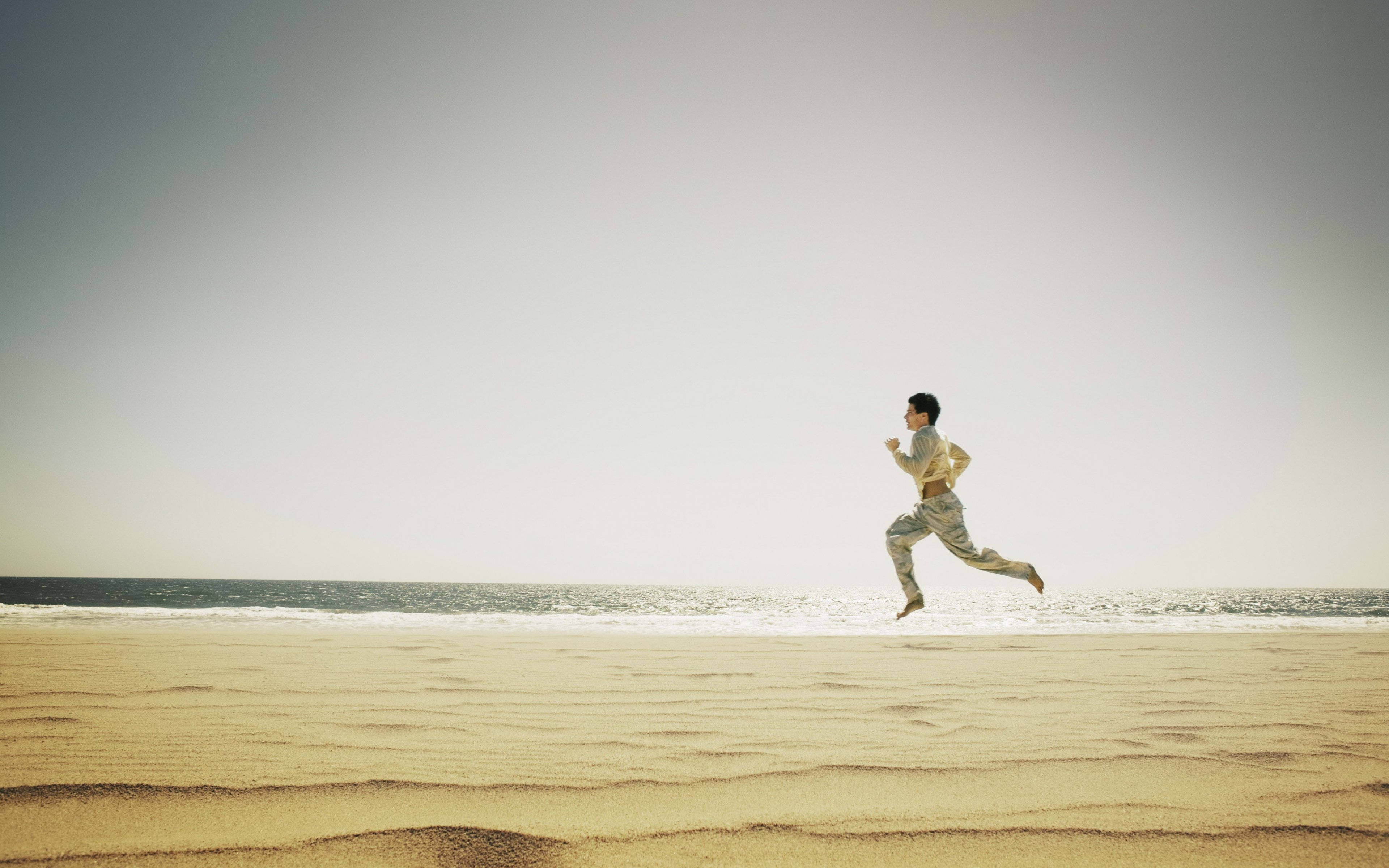 Running 桌布 壁紙 — 一個人在陽光普照的沙灘上赤腳奔跑，海浪在蒼白的天空下翻滾。