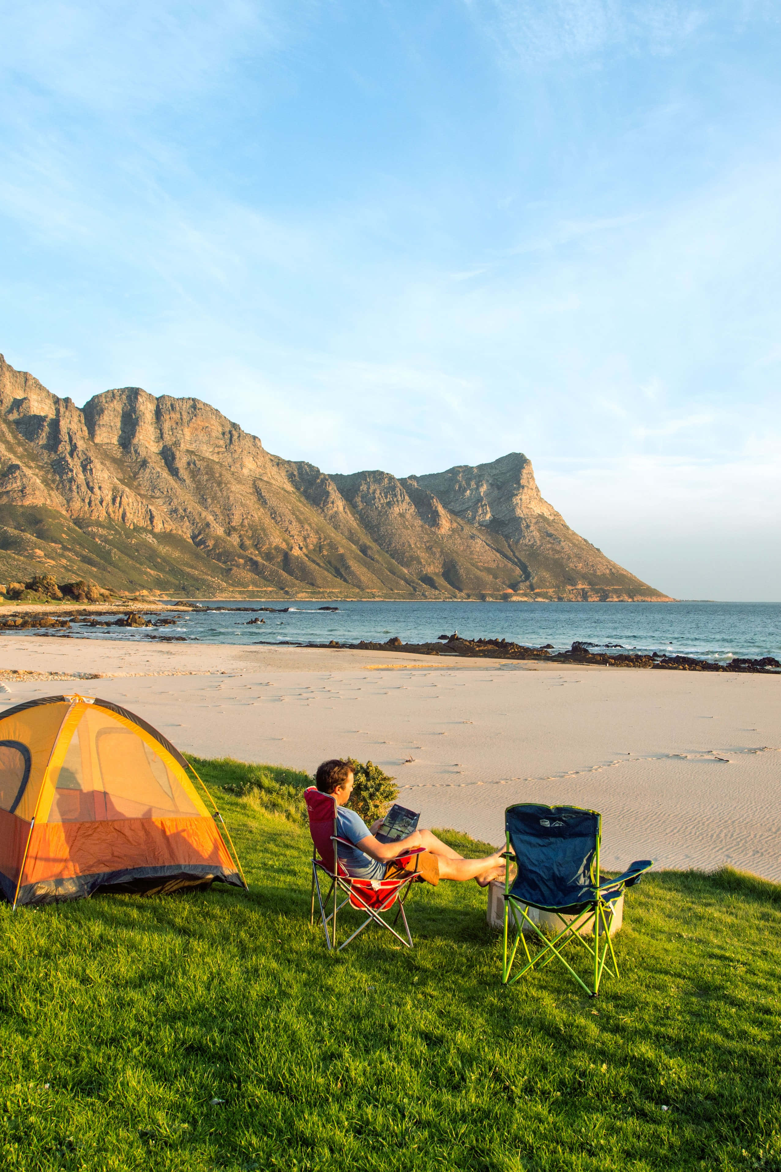 Beach Camping 桌布 壁紙 — 一個人在青草覆蓋的山坡上，坐在紅色椅子上放鬆，俯瞰沙灘和巍峨的山脈，天空清澈湛藍。