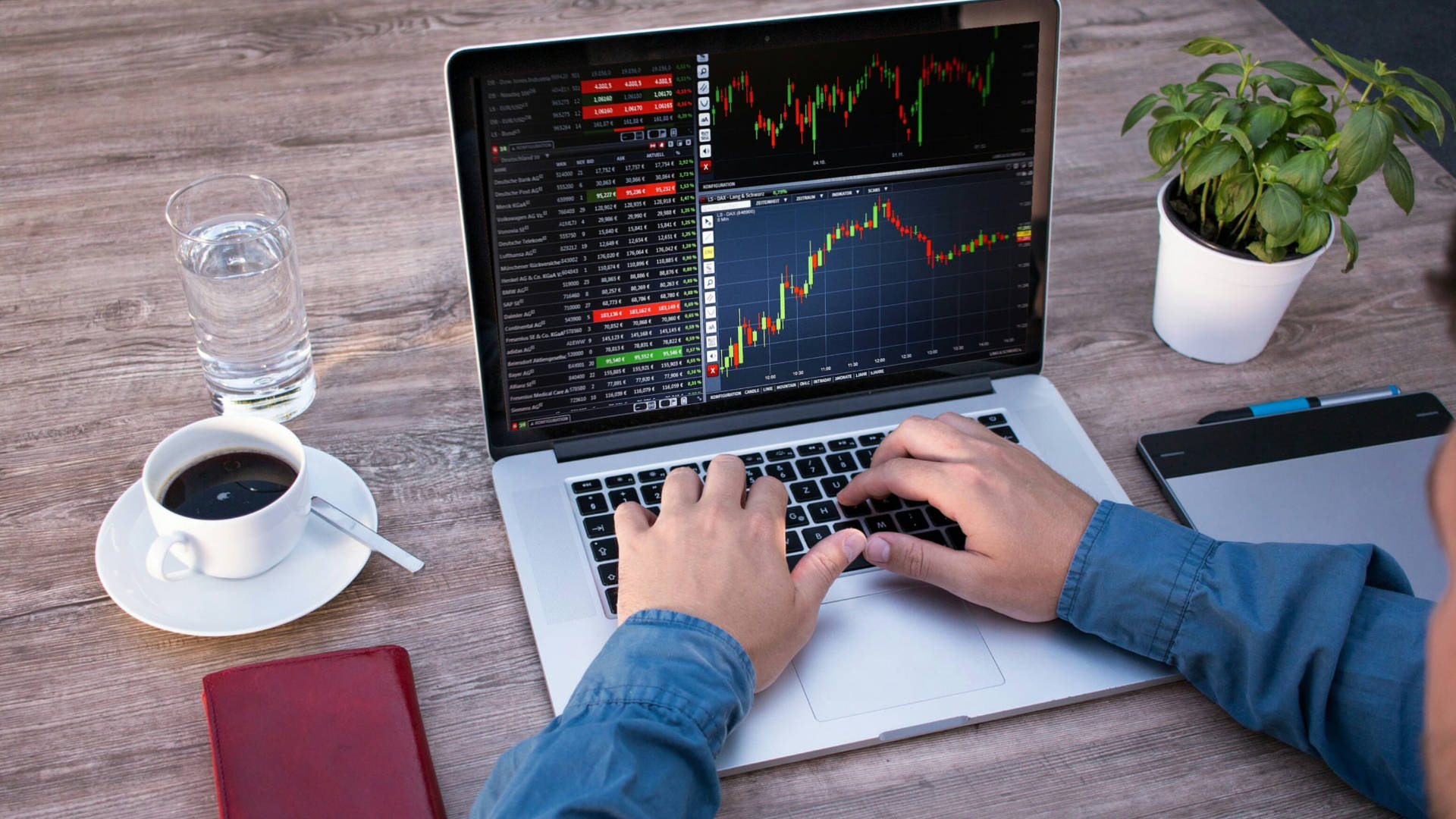 Forex 桌布 壁紙 — 一個人在顯示股市圖表的筆記型電腦上打字，木桌上放著咖啡和盆栽植物。