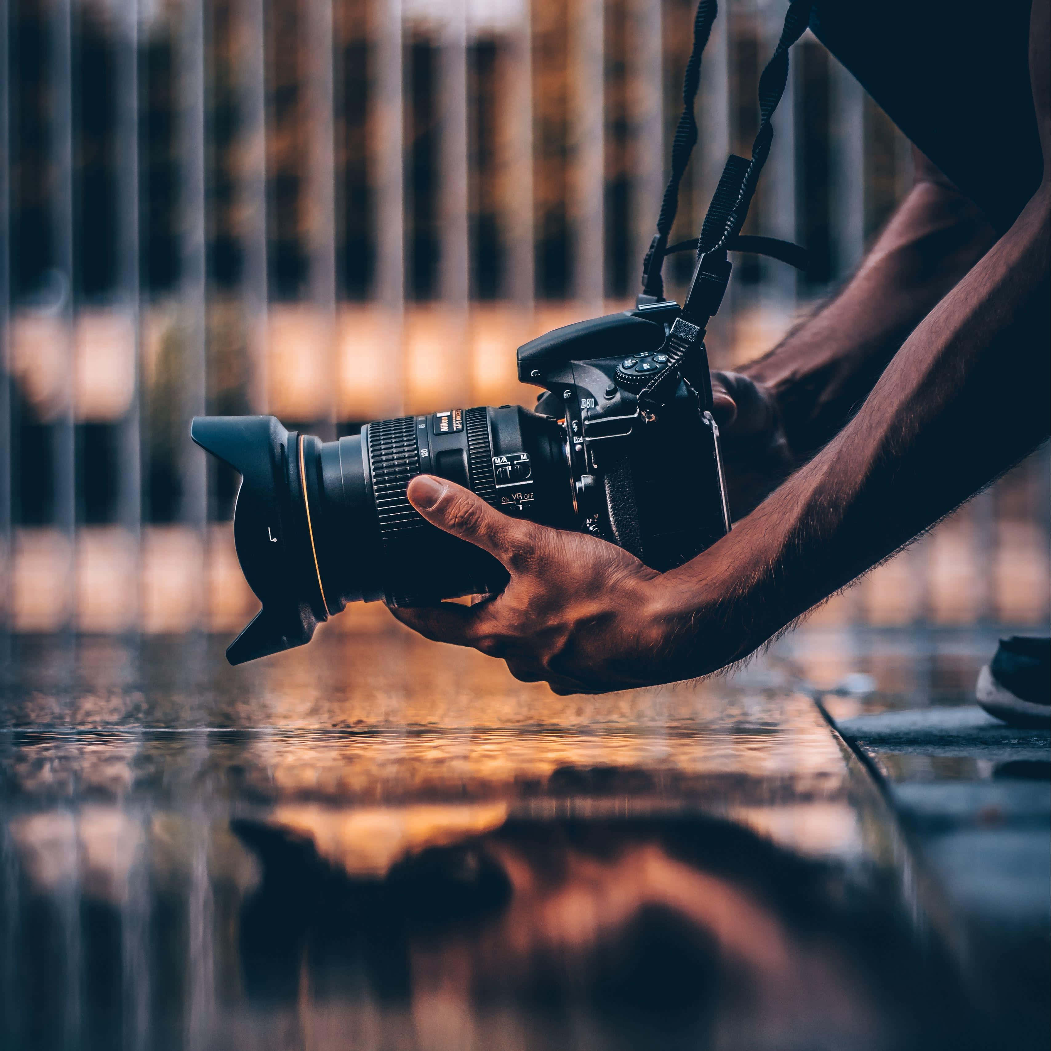 Dslr 桌布 壁紙 — 一個人在黃昏時分，手持DSLR相機，鏡頭對準被雨水打濕、反射著溫暖金色城市燈光的路面。