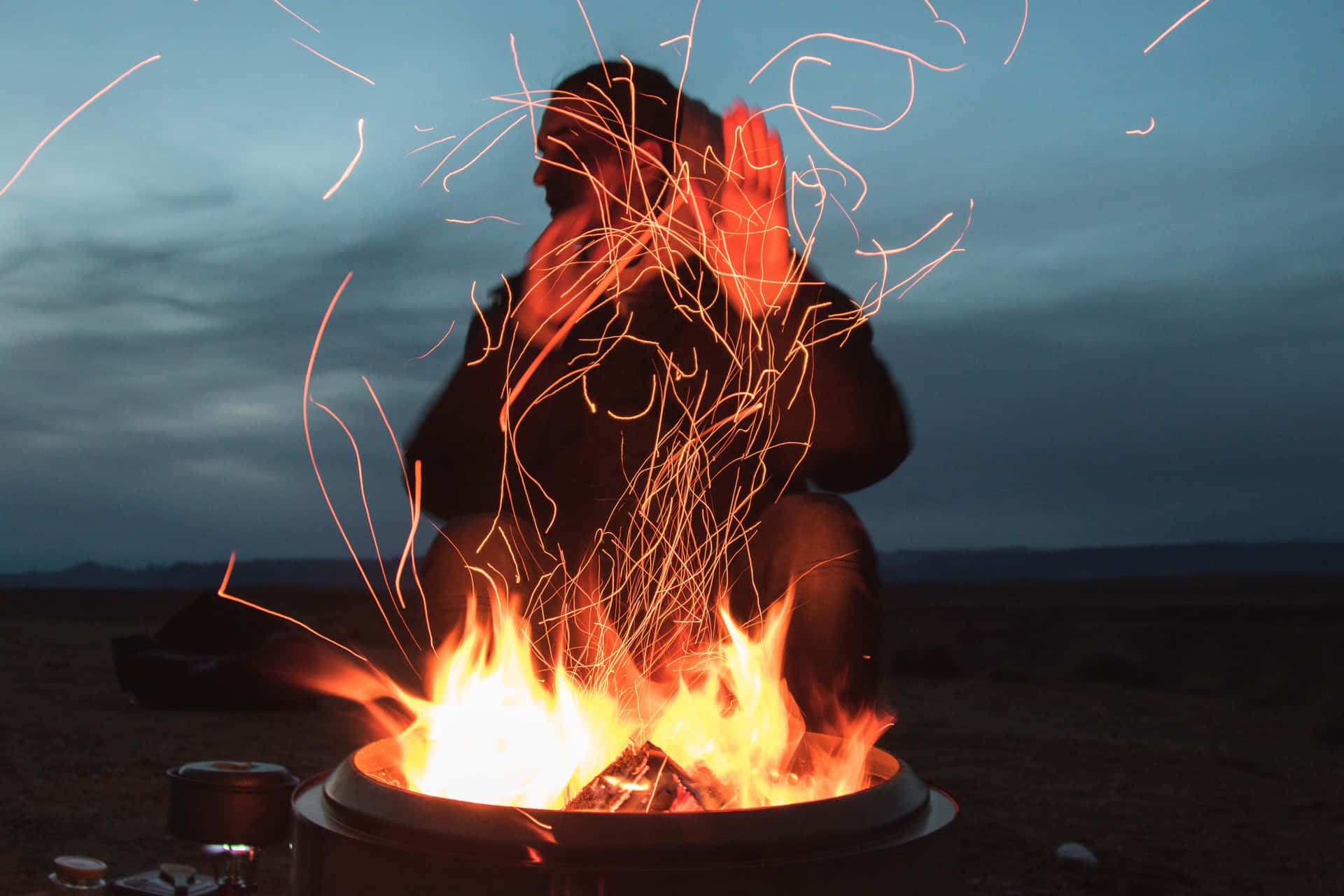 Fall Bonfire 桌布 壁紙 — 一個人在黃昏時分坐在熊熊燃燒的營火旁，火花向漸暗的天空飛舞。