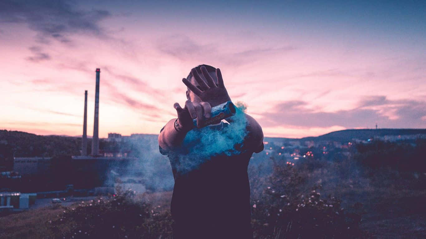 chill 桌布 壁紙 — 一個人在黃昏時分戶外，從罐中釋放藍色煙霧，背景是粉紅和紫色的天空，遠處有工業煙囪。