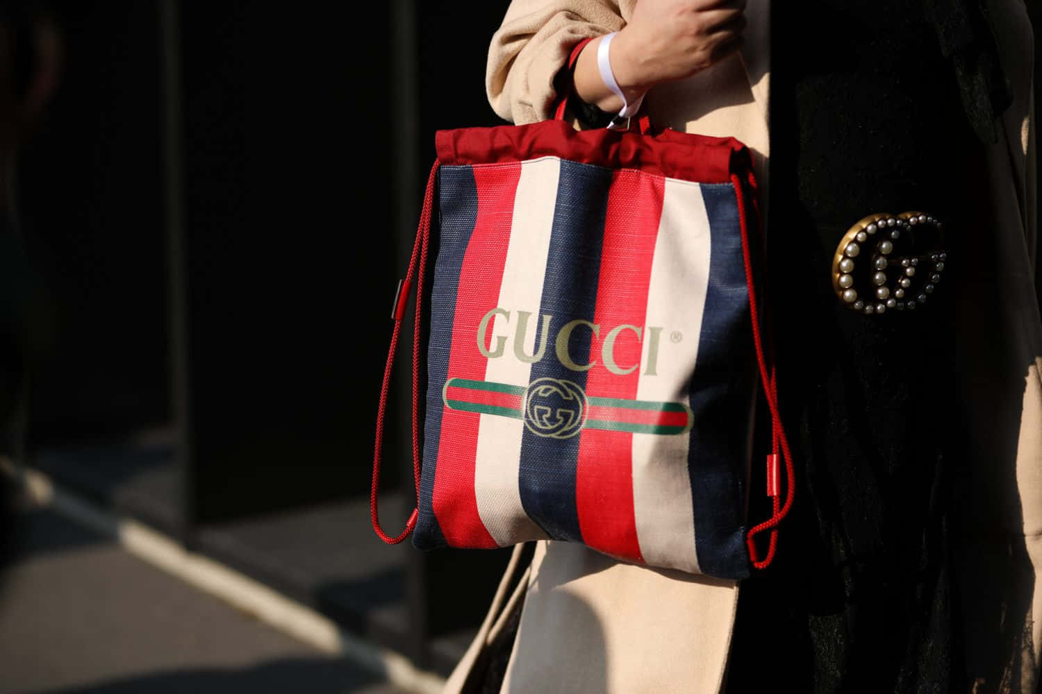 Gucci 桌布 壁紙 — 一個人在黑暗的城市背景下，拿著一個紅白藍條紋Gucci束繩袋，上面有金色標誌。