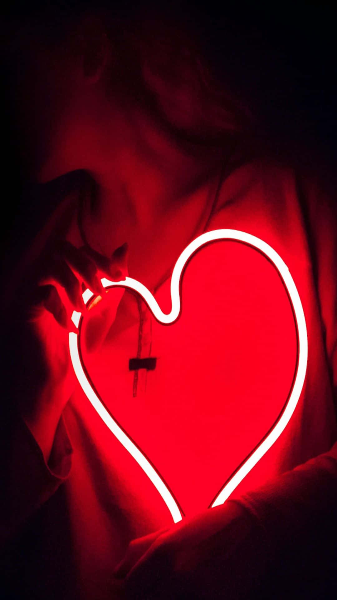 Neon Lights Aesthetic 桌布 壁紙 — 一個人在黑暗背景前，拿著一個發光的紅色霓虹心形，在他們的臉和手上投射出溫暖的紅光。