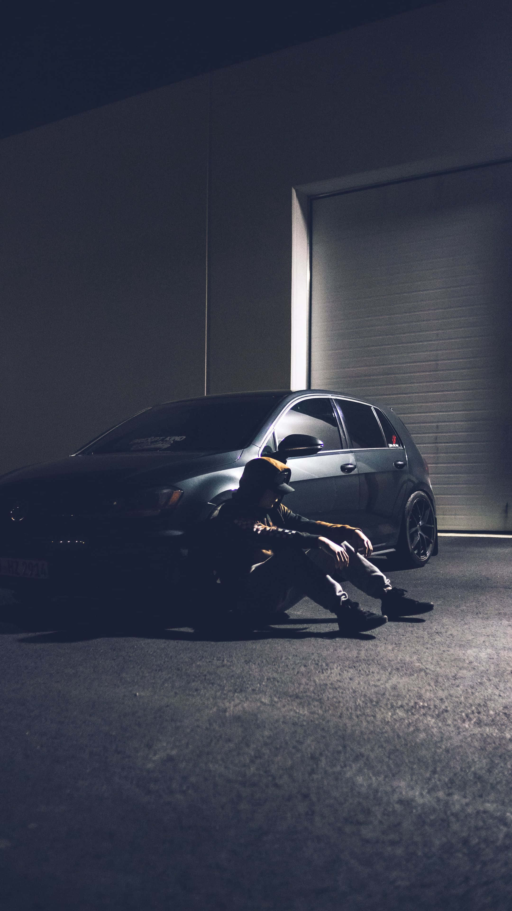 Sad Vibes 桌布 壁紙 — 一個人坐在一輛深色汽車旁邊，車庫光線昏暗，只有一道光束照亮場景。