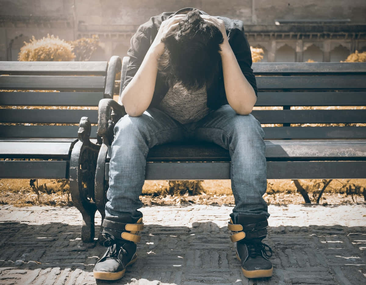 Depressed 桌布 壁紙 — 一個人坐在公園長椅上，雙手抱頭，在溫暖的陽光下流露出深深的痛苦。
