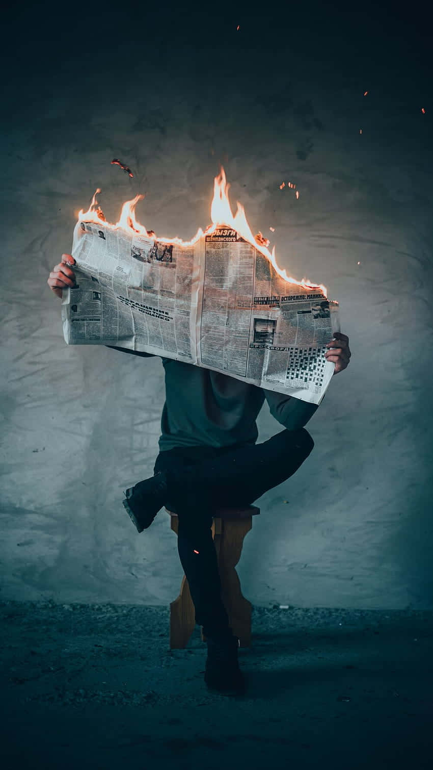 Newspaper 桌布 壁紙 — 一個人坐在凳子上，手持燃燒的報紙遮住臉部，背景是深色紋理牆壁。