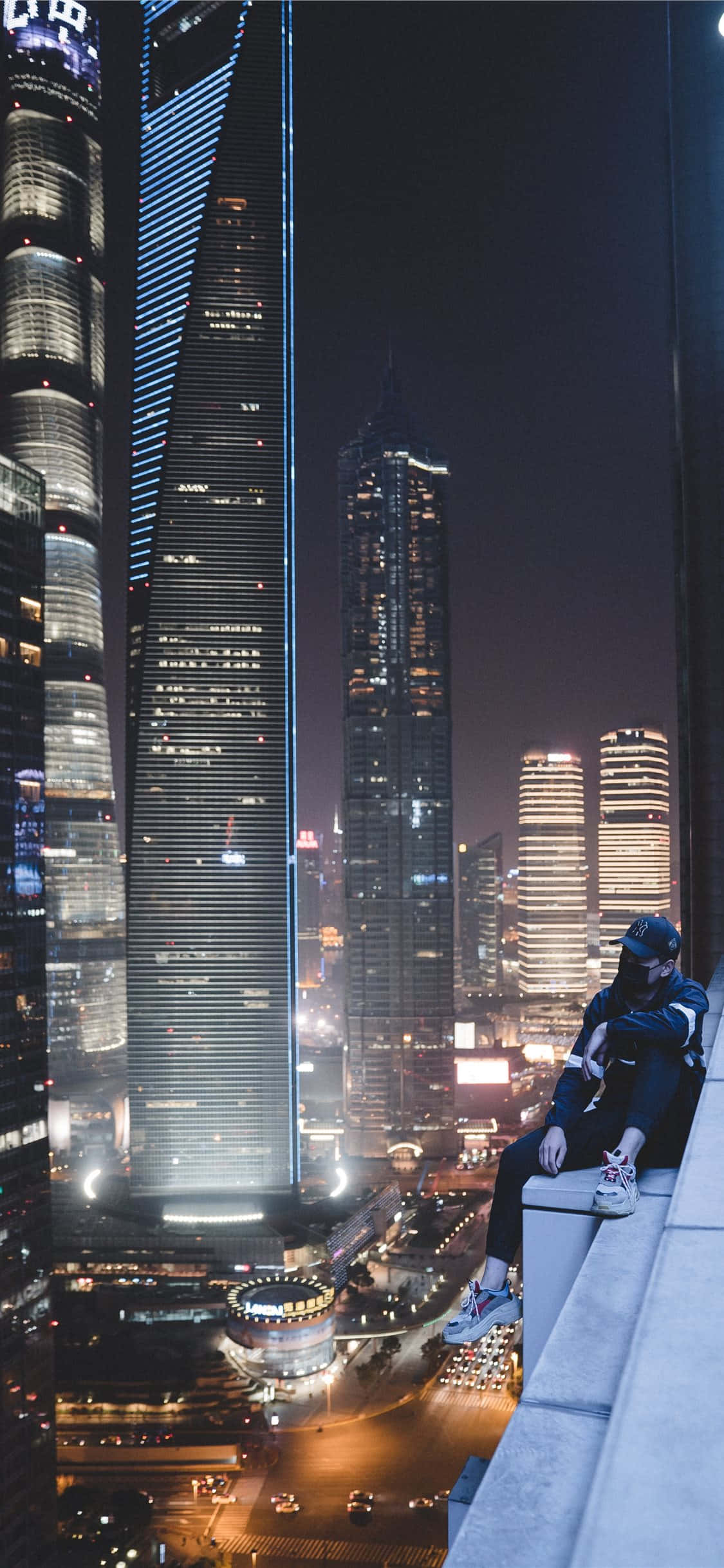 Rooftop 桌布 壁紙 — 一個人坐在屋頂邊緣，俯瞰下方閃爍著燈光的摩天大樓和明亮街道構成的迷人夜景。