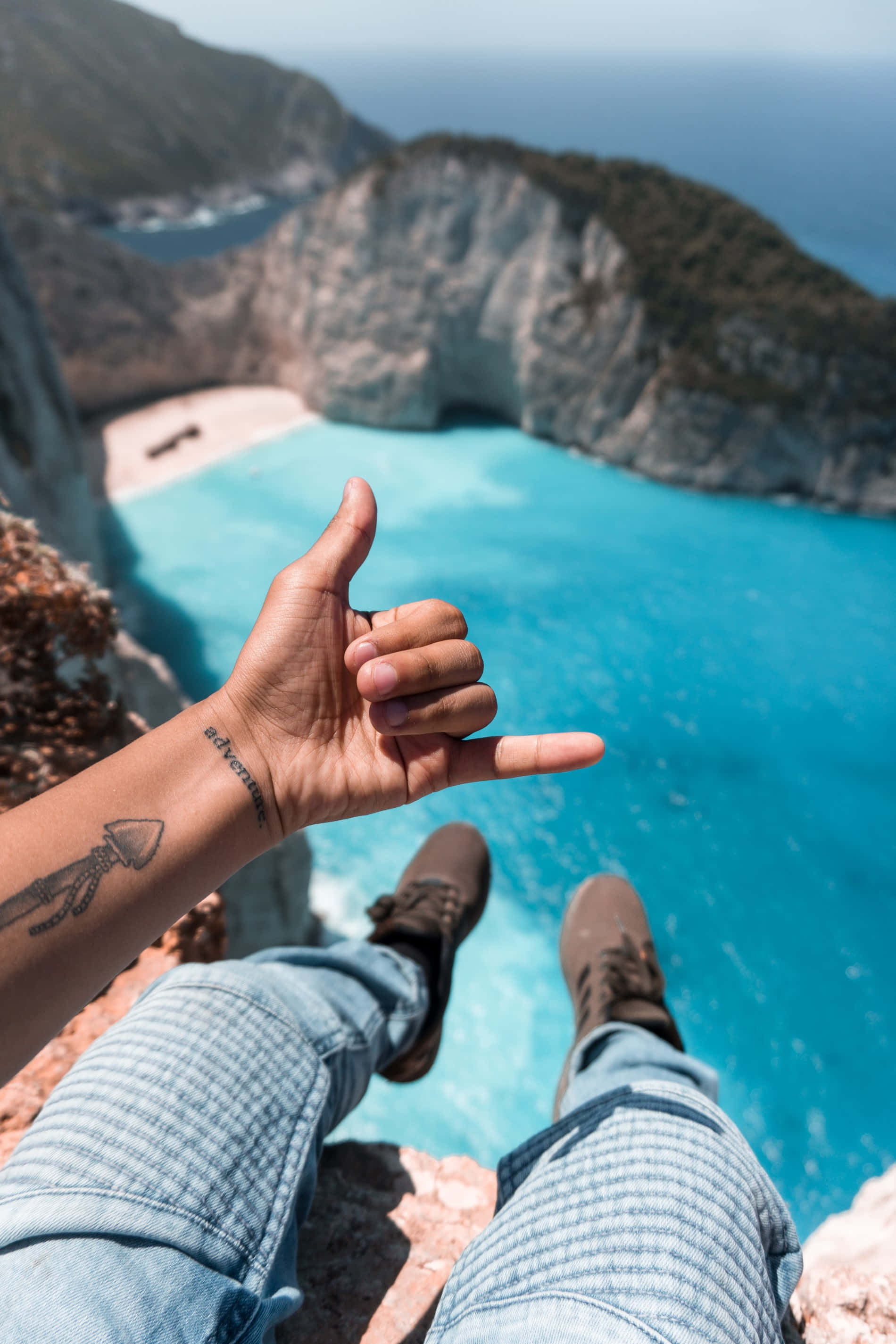 Travel Aesthetic Phone 桌布 壁紙 — 一個人坐在懸崖上，俯瞰著碧綠的海灣，舉起大拇指，露出刺青的手臂和牛仔褲。