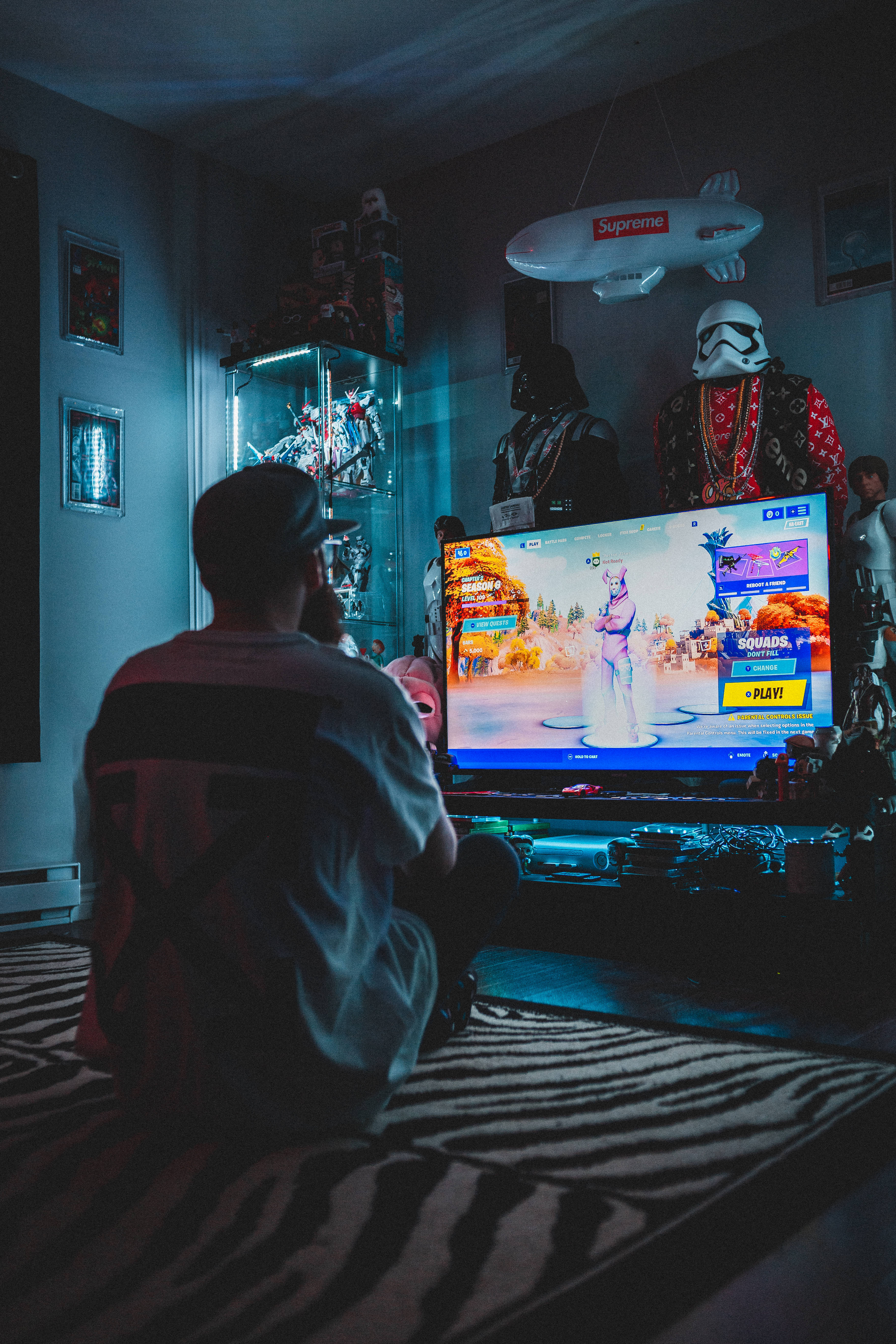 遊戲 桌布 壁紙 — 一個人坐在斑馬紋地毯上，在昏暗的房間裡看著發光的電視播放Fortnite遊戲，周圍是流行文化紀念品。
