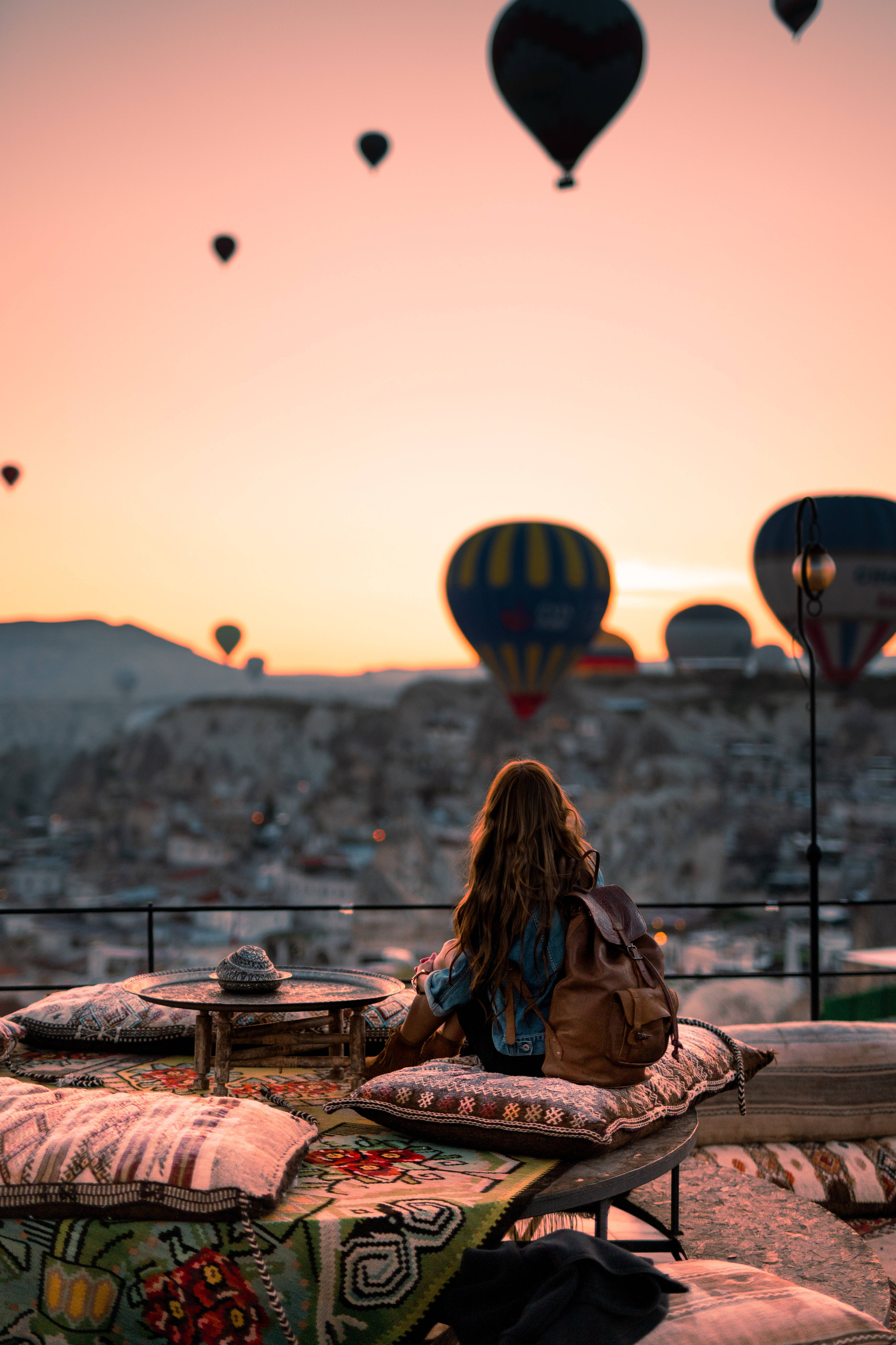 Cappadocia 桌布 壁紙 — 一個人坐在有圖案的坐墊上，俯瞰山谷，熱氣球在日落時升起。