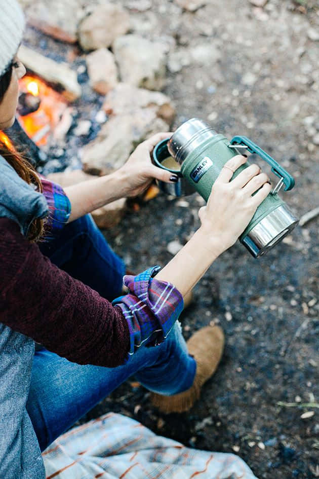 Minimalist Camping 桌布 壁紙 — 一個人坐在營火旁，手持綠色保溫瓶，戴著毛帽和格紋襯衫。