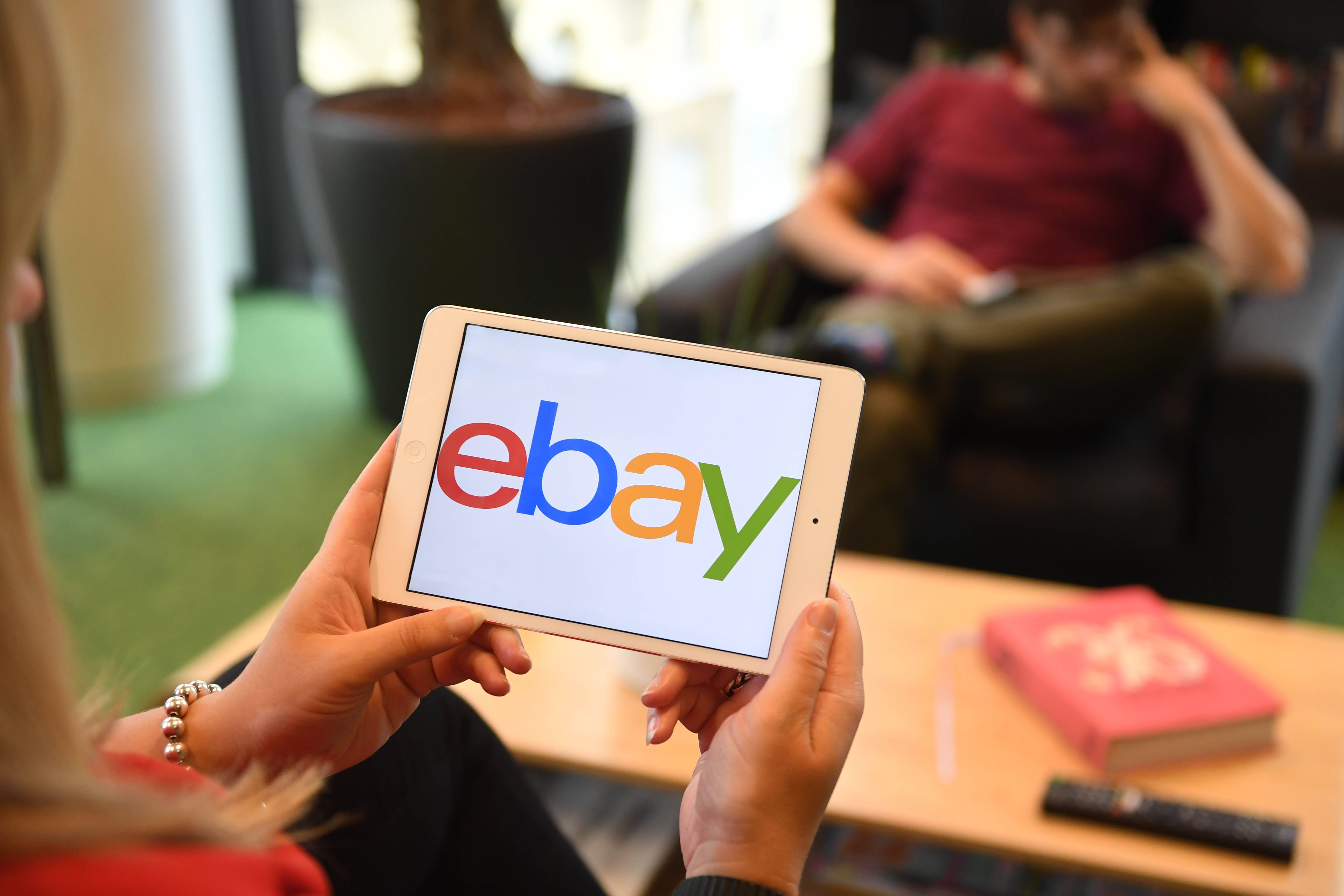 eBay 桌布 壁紙 — 一個人坐在現代客廳中，手持顯示eBay標誌的平板電腦，背景有另一個人。