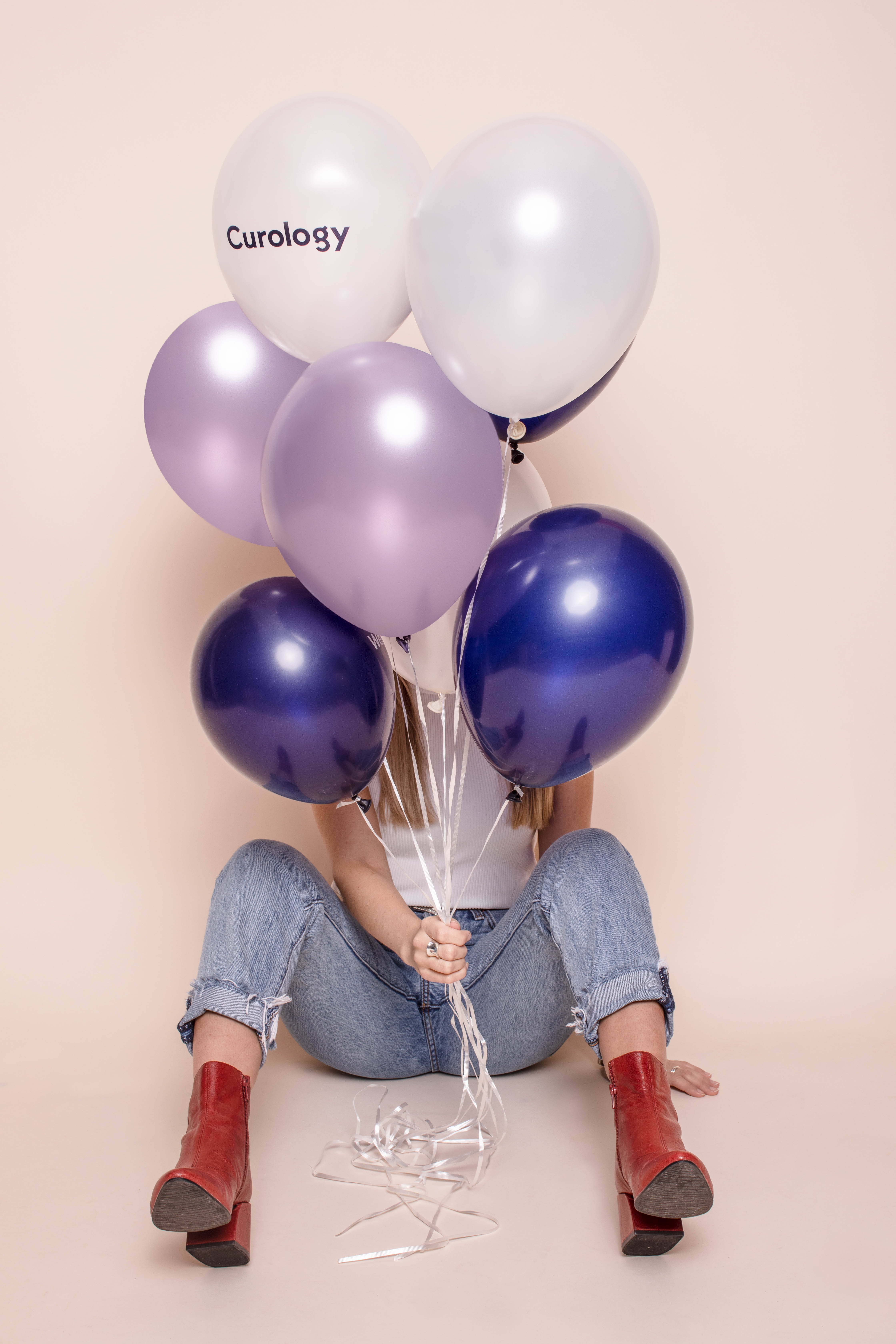 Balloons 桌布 壁紙 — 一個人坐在米色地板上，拿著一束閃亮的紫色、藍色和白色氣球，其中一個寫著 Curology。