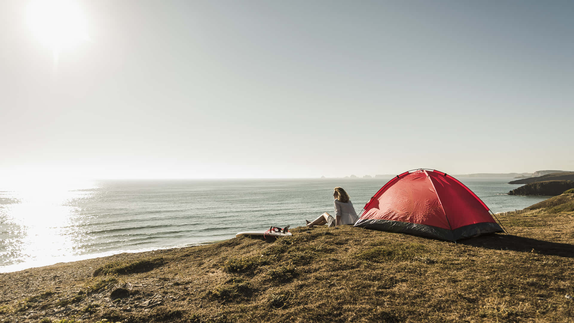 Beach Camping 桌布 壁紙 — 一個人坐在草地的懸崖邊，旁邊有一個紅色帳篷，俯瞰陽光普照的海洋和遠處島嶼。