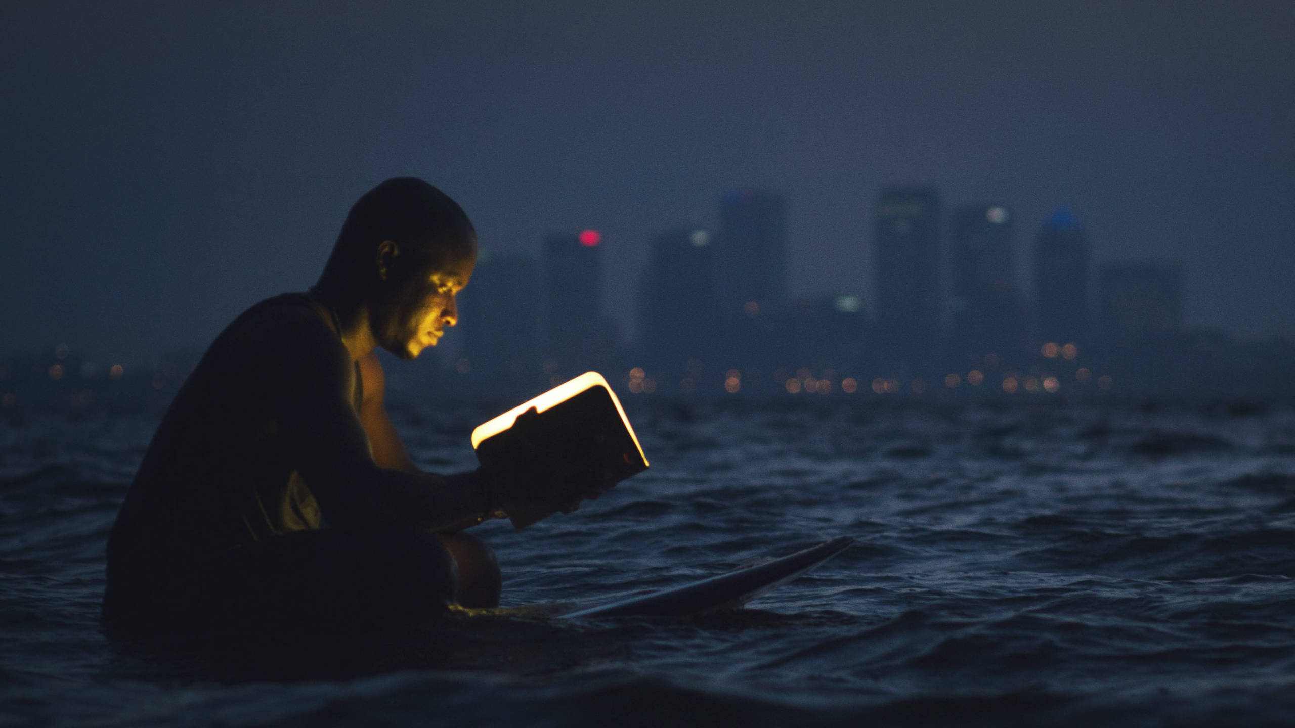 Reading 桌布 壁紙 — 一個人夜晚坐在黑暗的水中，閱讀一本發光的書，背景是模糊的城市天際線。