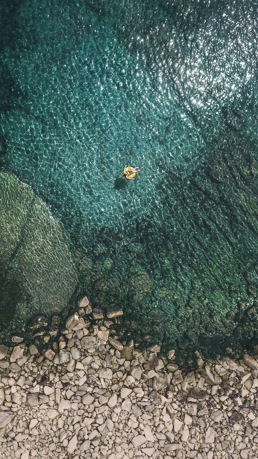 iOS 11 桌布 壁紙 — 一個人孤獨地漂浮在明亮的黃色充氣圈上，在岩石海岸附近清澈的碧綠海水中，寧靜祥和。
