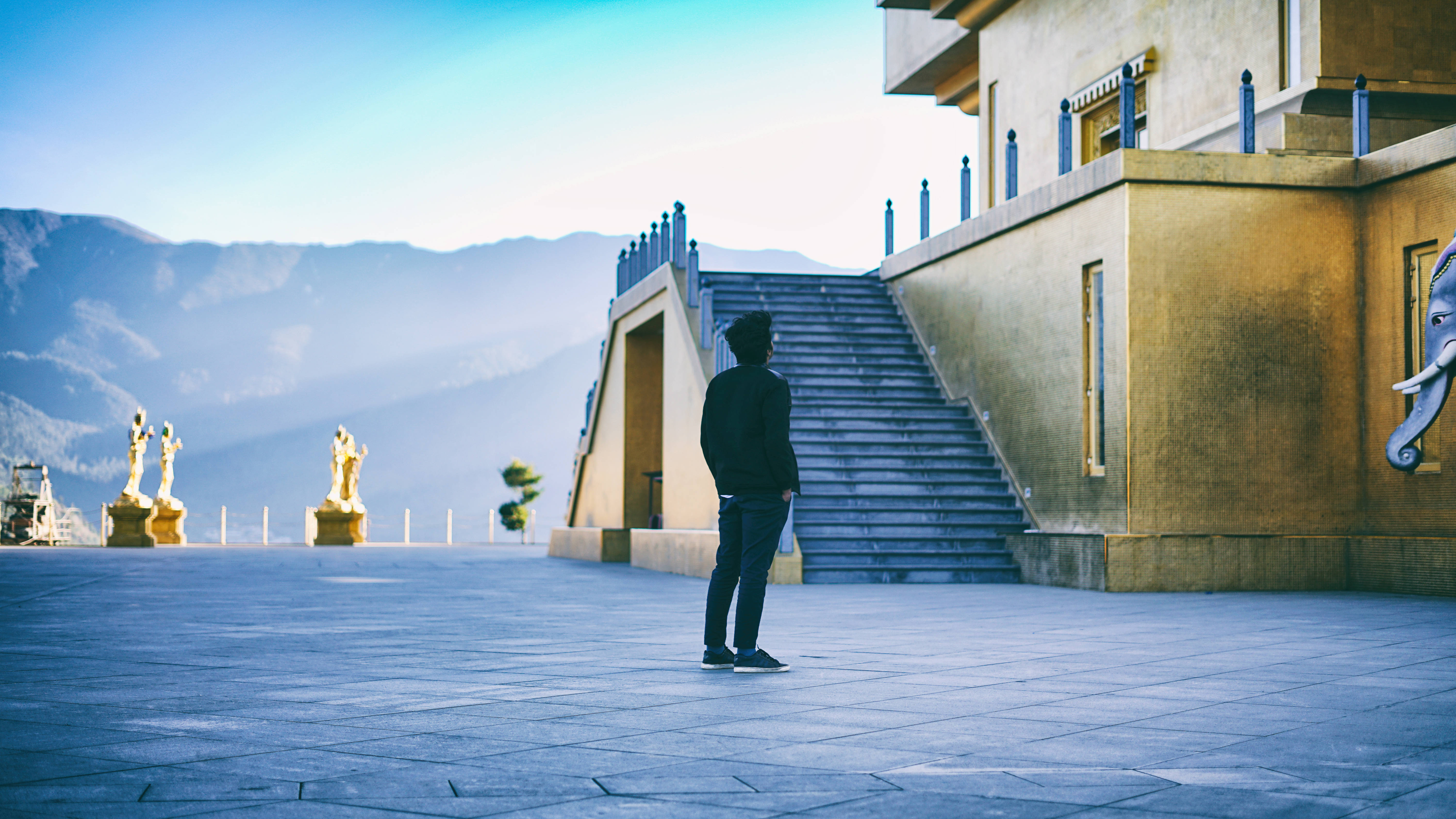 Bhutan 桌布 壁紙 — 一個人孤獨地站在瓷磚廣場上，面前是一座宏偉的寺廟，有金色雕像和遠山，在清澈的藍天下。