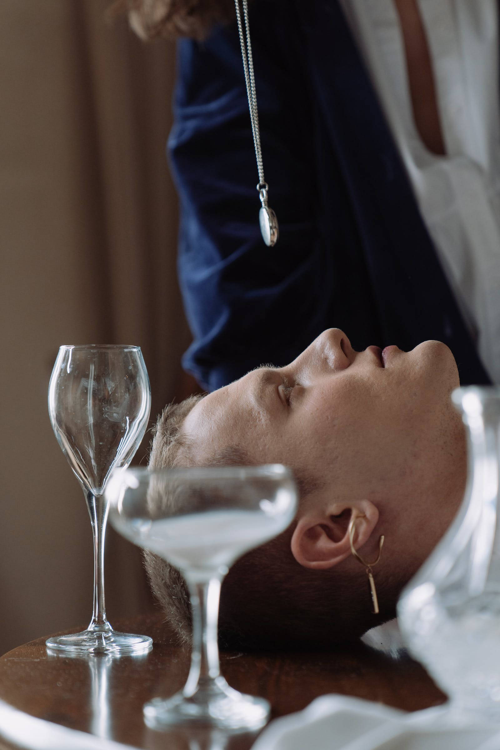 Hypnosis 桌布 壁紙 — 一個人將頭靠在木桌上，旁邊有空的酒杯，上方懸掛著一條吊墜項鍊。