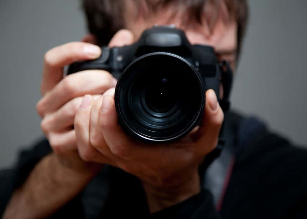 Holding Camera 桌布 壁紙 — 一個人將黑色DSLR相機舉到臉前，背景為純灰色。