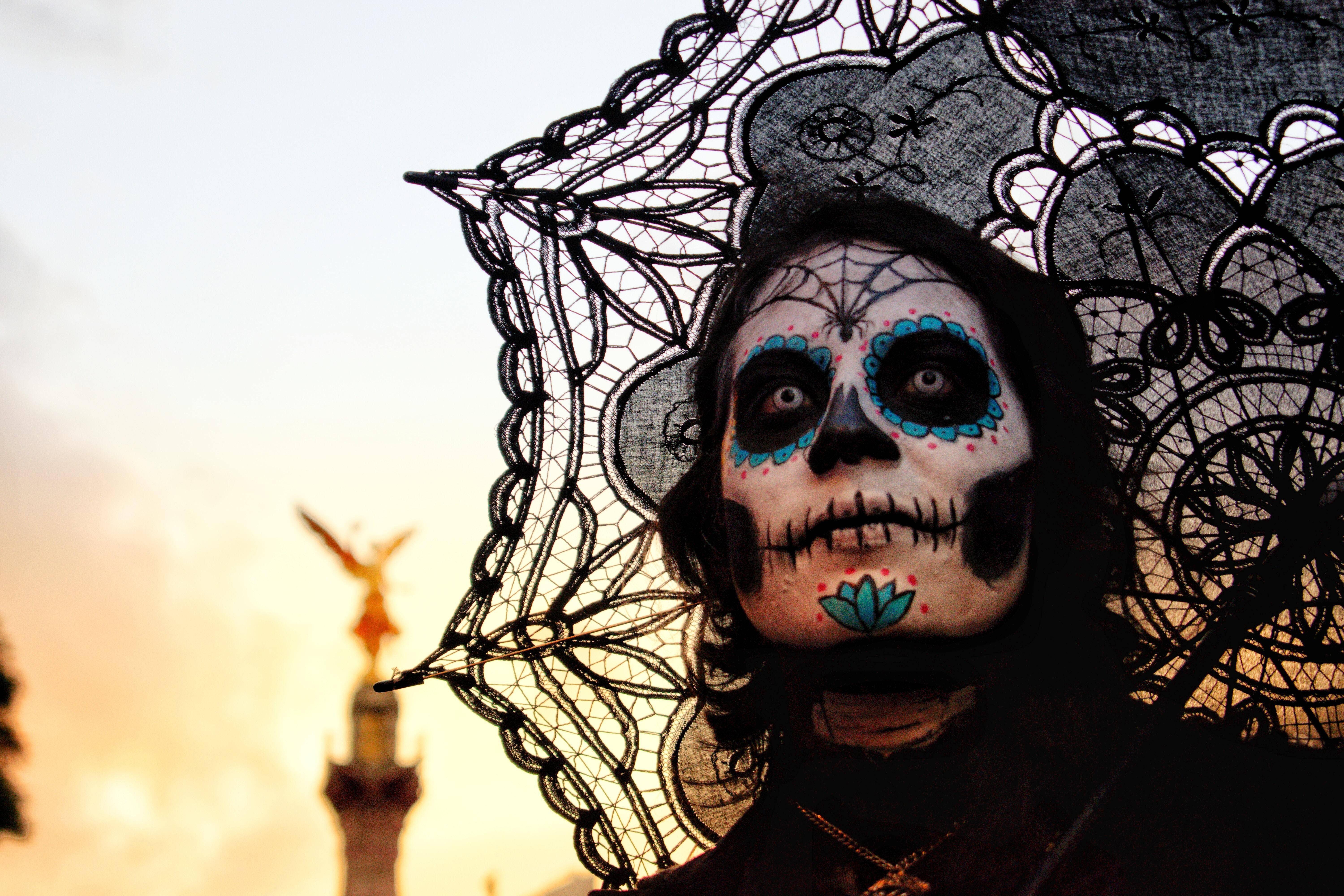 Dia De Los Muertos 桌布 壁紙 — 一個人帶著精緻的糖骷髏妝容和蕾絲陽傘，站在溫暖的日落背景下，遠處有一座金色雕像。
