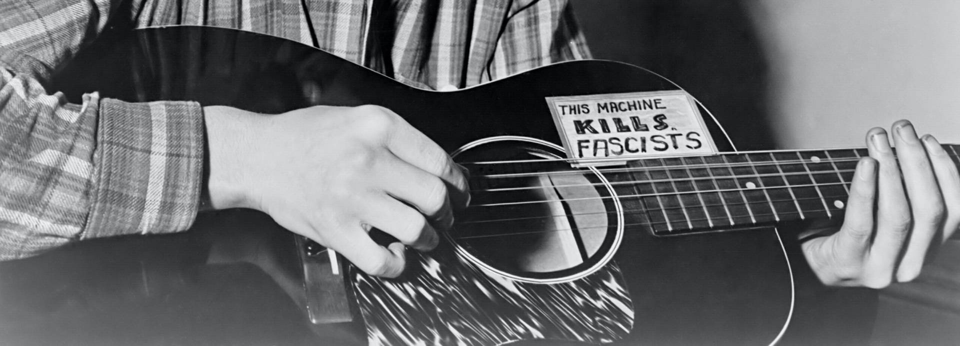 黑白 桌布 壁紙 — 一個人彈奏木吉他，吉他身上貼有寫著「THIS MACHINE KILLS FASCISTS」的大膽貼紙。