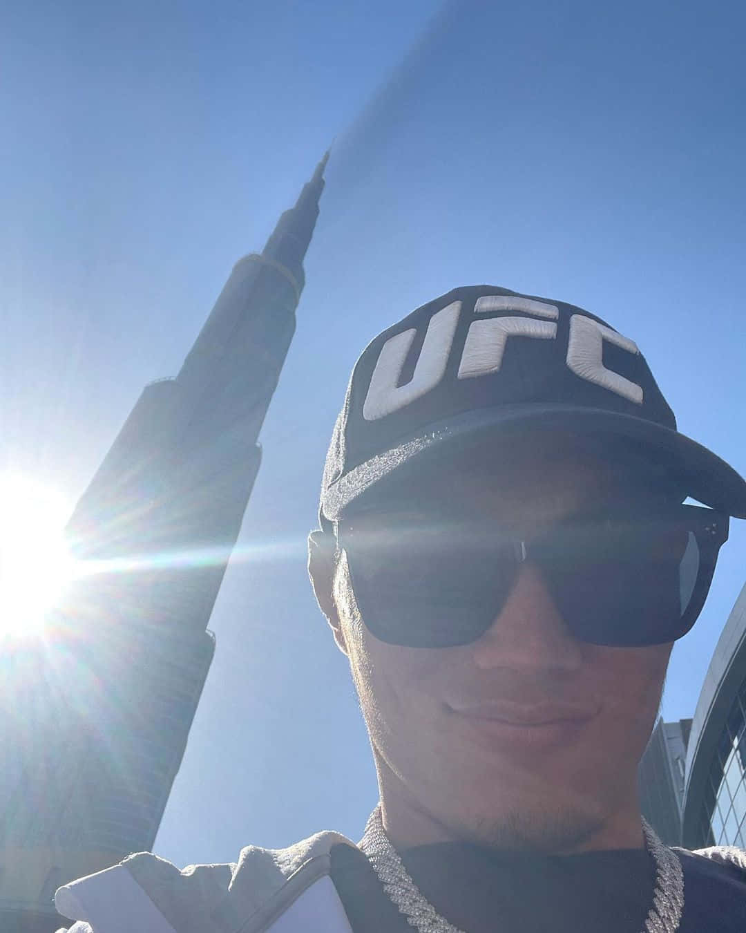 Sun Rays 桌布 壁紙 — 一個人戴著UFC帽子和太陽眼鏡，在明亮的陽光下於哈里發塔前面微笑。