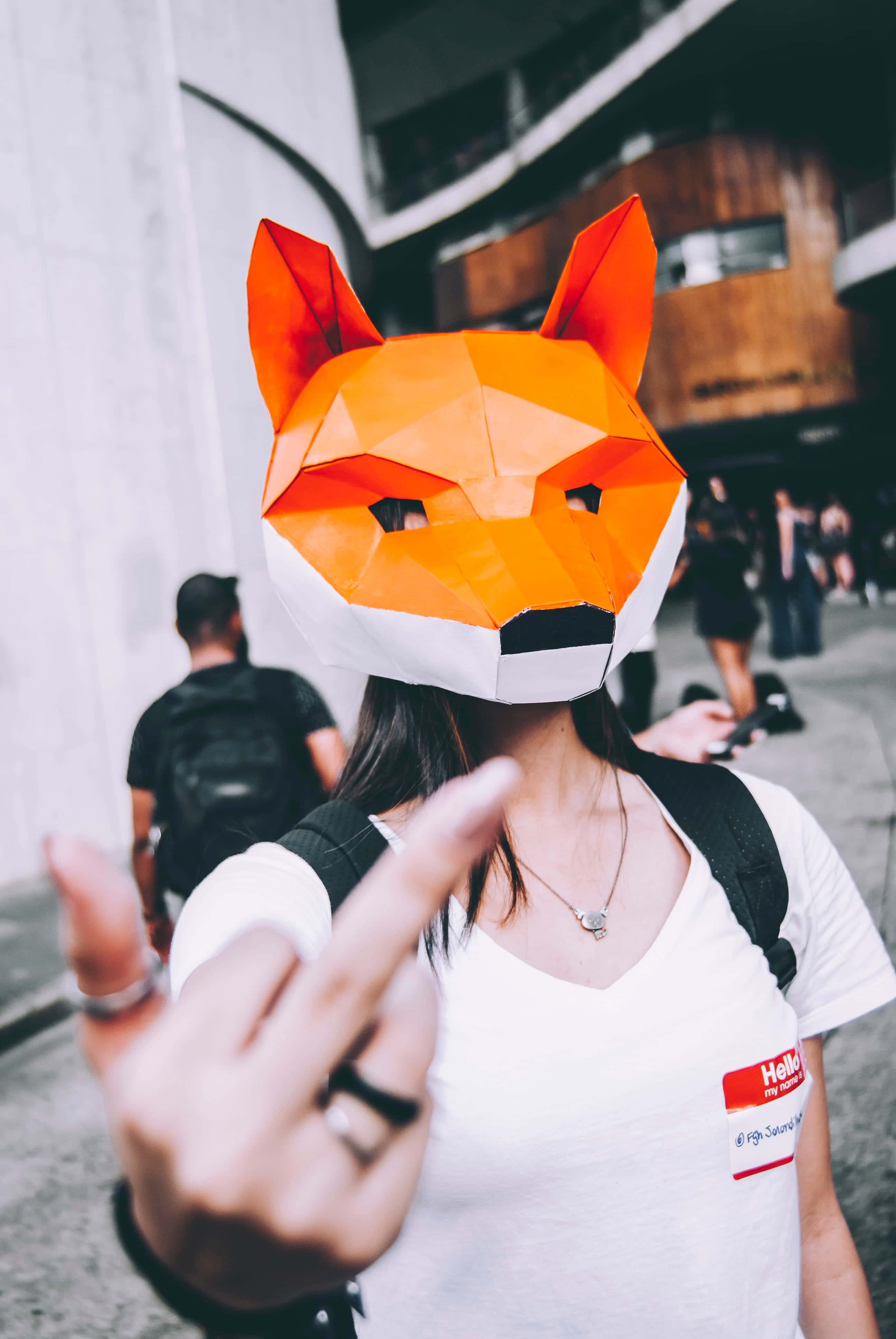 rude 桌布 壁紙 — 一個人戴著亮橙色幾何狐狸面具，在繁華的城市街道上直接指向相機。