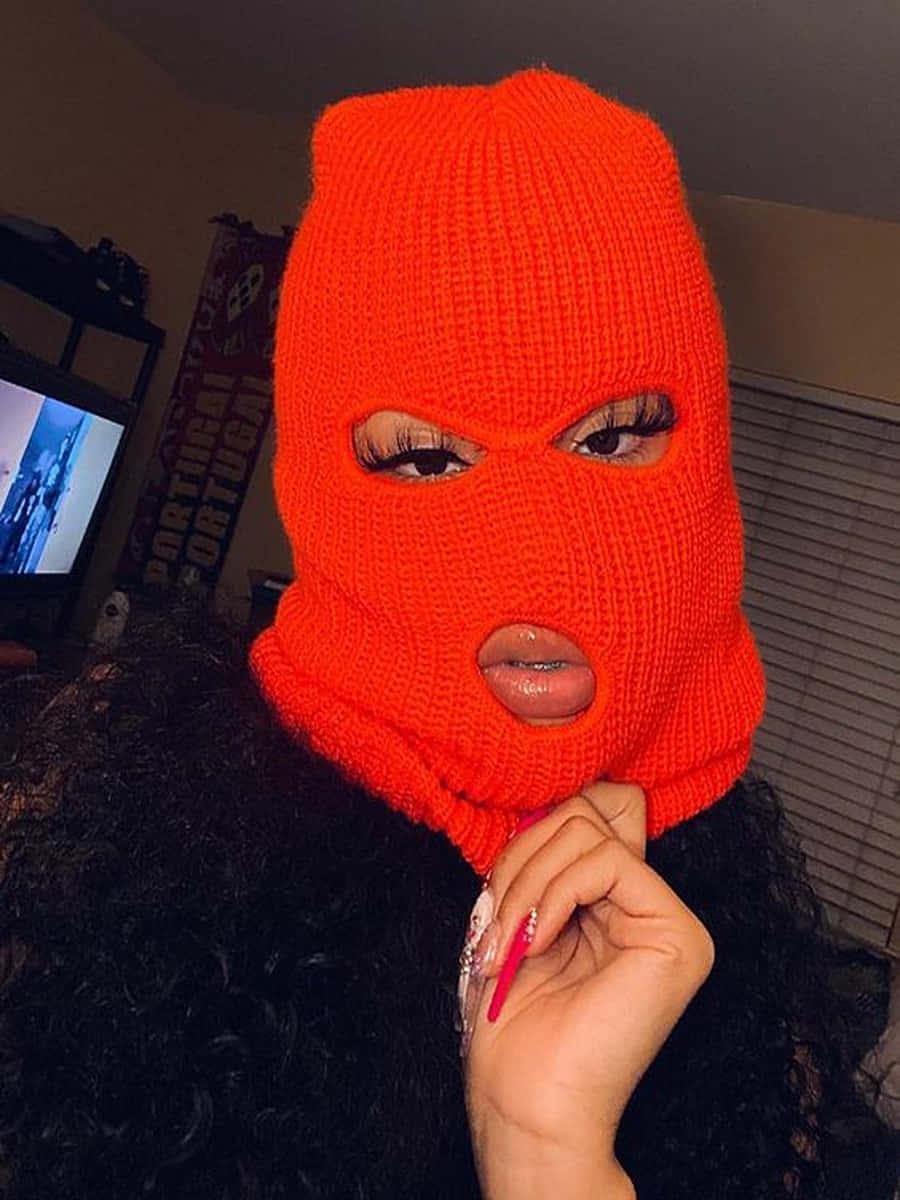 Ski Mask Girl 桌布 壁紙 — 一個人戴著亮橙色針織巴拉克拉法帽，只露出眼睛和嘴巴，在下巴附近拿著一支粉紅色筆。
