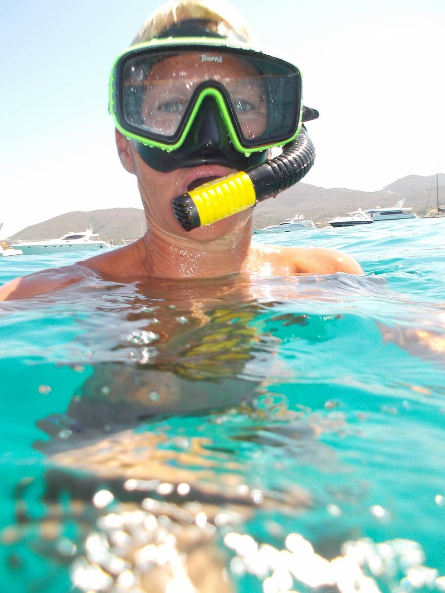 Snorkeling 桌布 壁紙 — 一個人戴著亮綠色潛水鏡和黃色呼吸管，從藍綠色海水中浮出，背景有船隻和山丘。