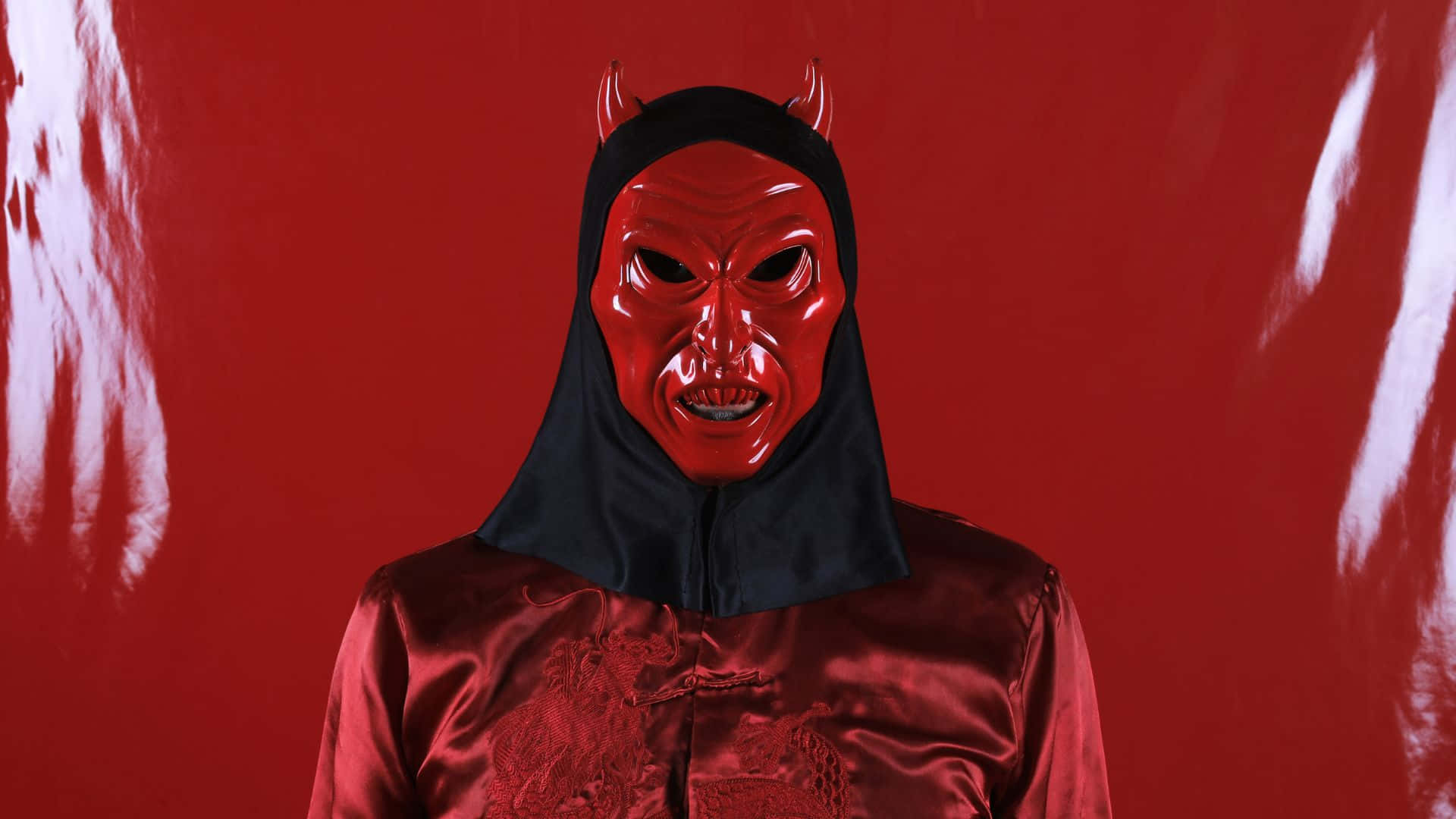 Horror Masks 桌布 壁紙 — 一個人戴著亮面紅色惡魔面具，有角及黑色頭罩，站在有白色油漆條紋的鮮紅色背景前。