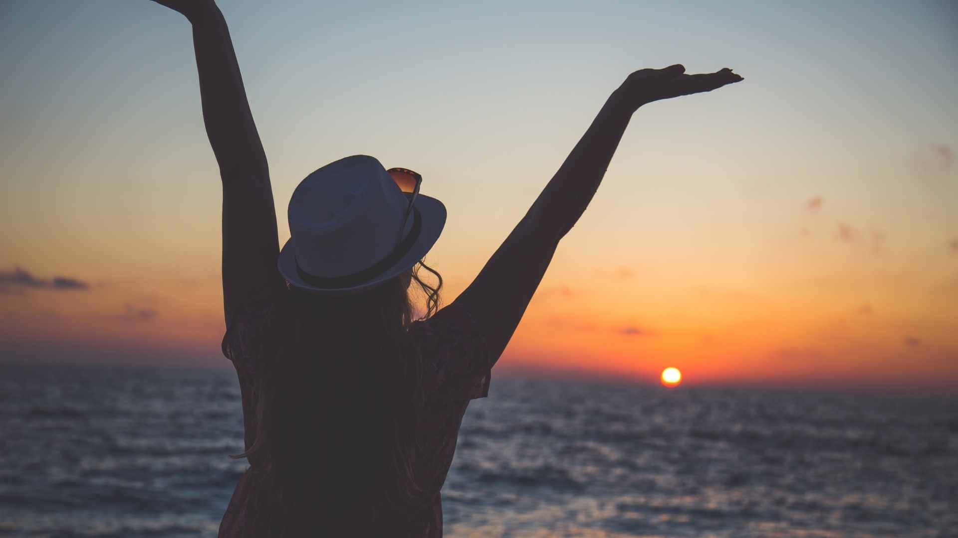 life 桌布 壁紙 — 一個人戴著帽子，在海洋上充滿活力的日落背景下，高舉雙臂，以喜悅的自由姿態呈現剪影。