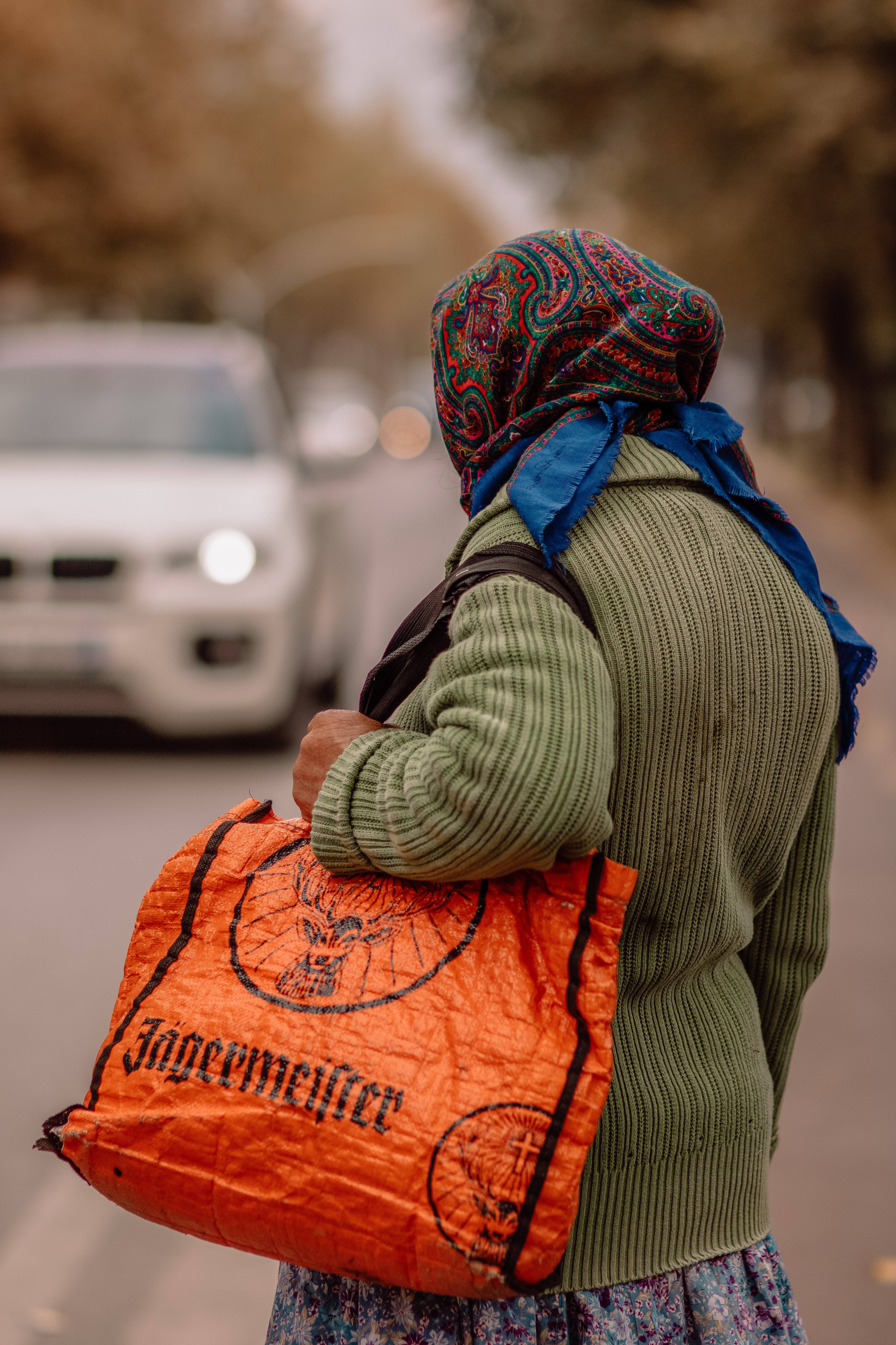 Albania 桌布 壁紙 — 一個人戴著彩色頭巾、穿著綠色毛衣，背著橙色Jägermeister袋，沿著街道行走，背景有一輛白色汽車。